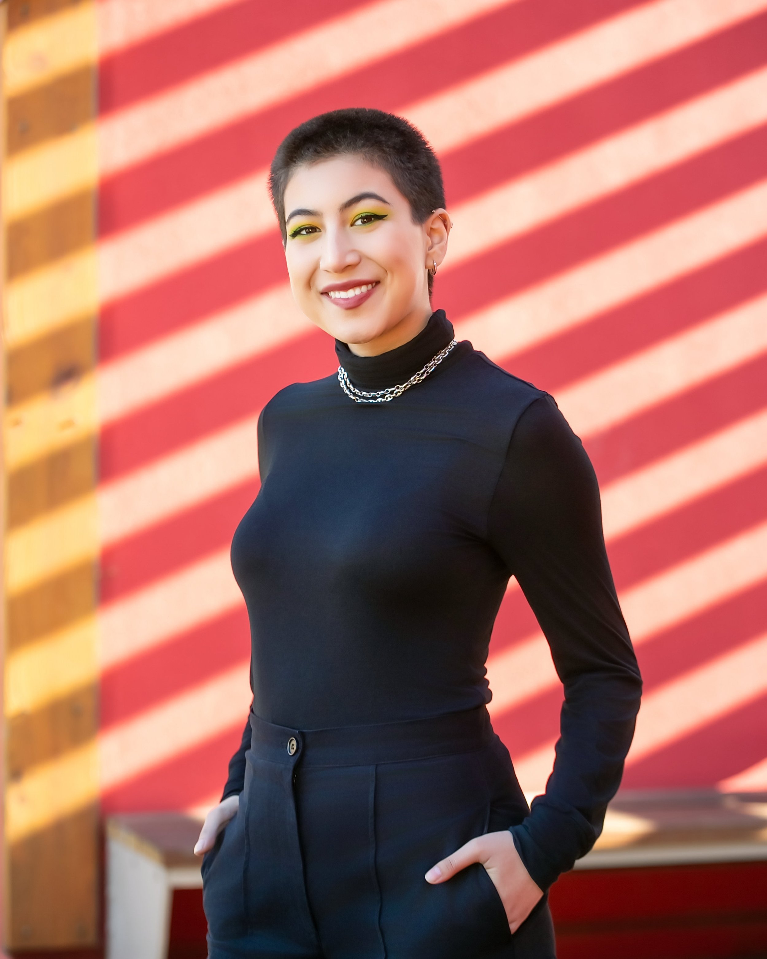 Portrait of a young woman with short dark hair, wearing a black turtleneck and layered silver necklaces, smiling, with a background of American flag.