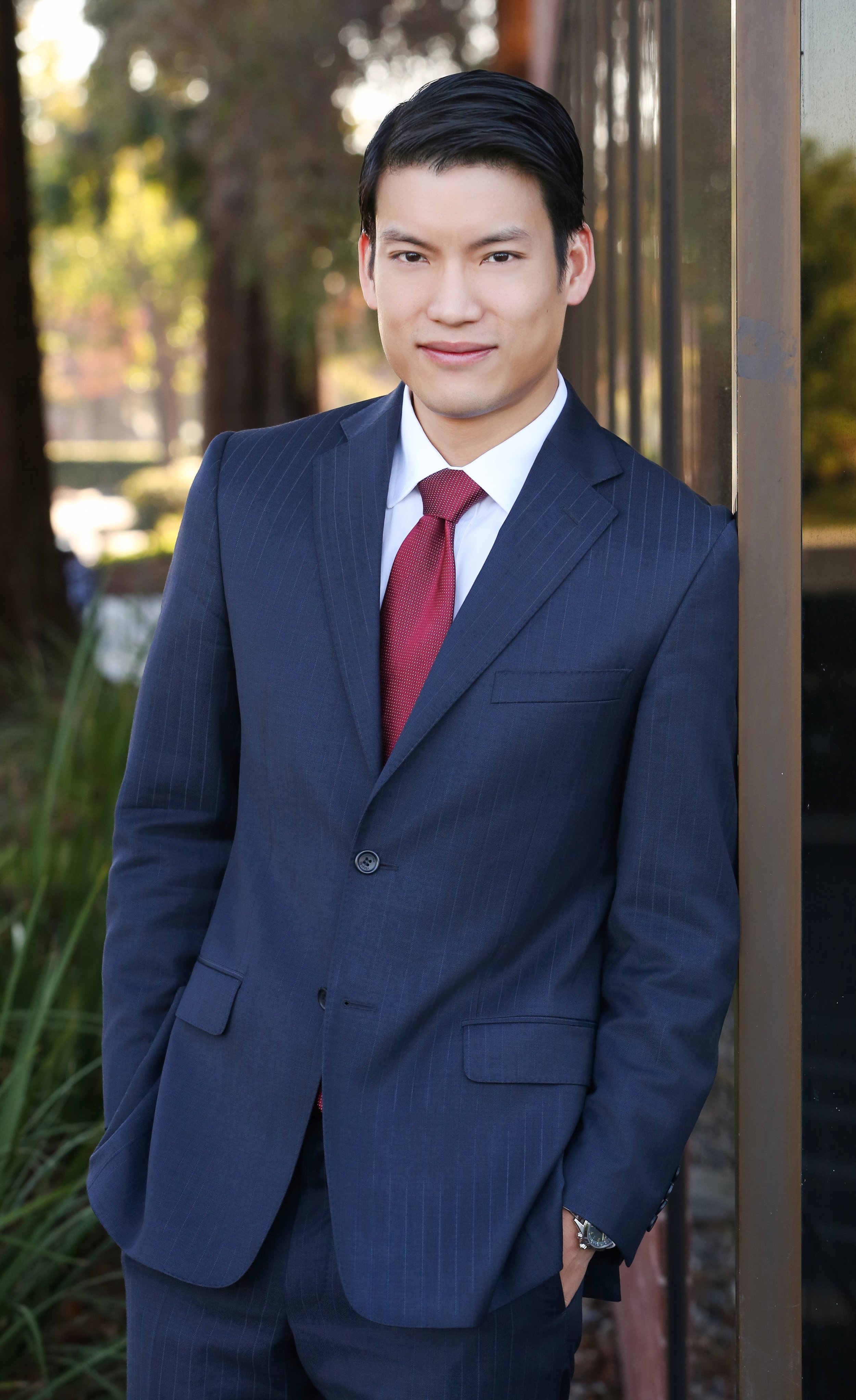 A man in a dark blue pinstripe suit with a white shirt and red tie standing outdoors, leaning against a corporate building with trees and greenery in the background.