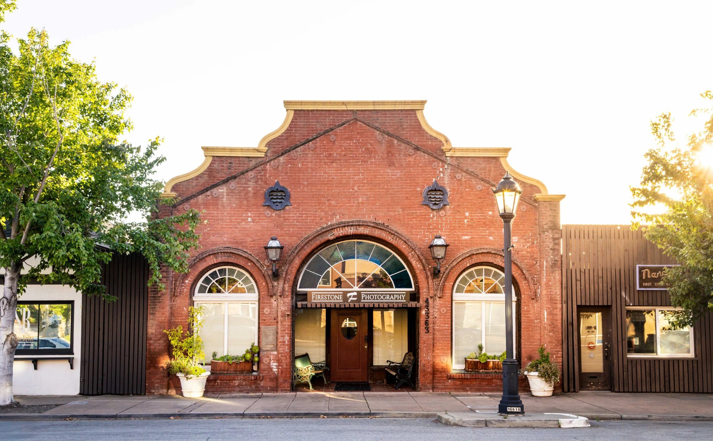 Front view of a historic red brick building with large arched windows and a sign that reads 'Firestone Photography.' The building has decorative brickwork and two vintage-style lamps flanking the entrance. There are trees and a streetlamp in the foreground, with the sun shining from the right side.