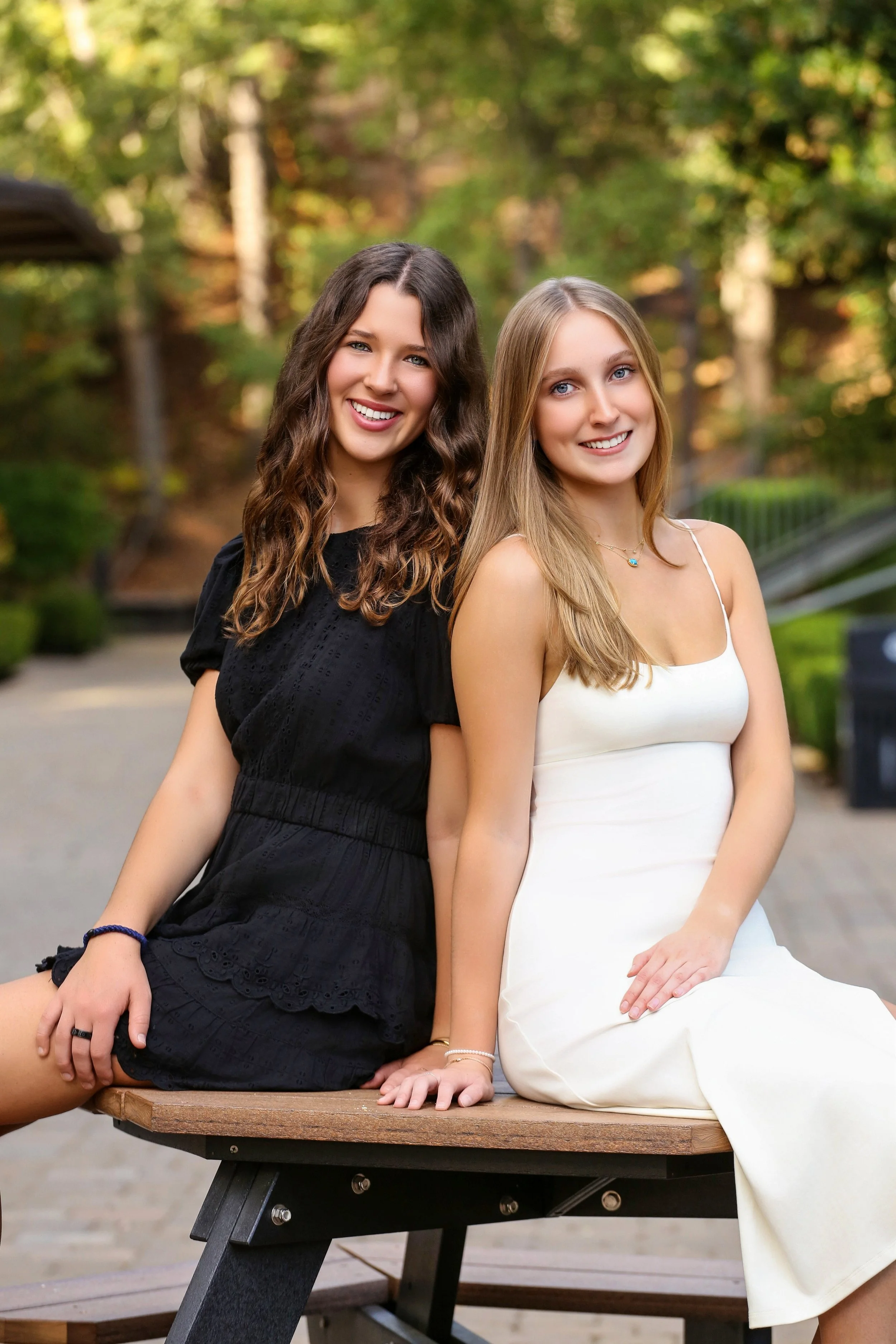 Two young women sitting on a bench outdoors, smiling at the camera, with a background of green trees and sunlight.