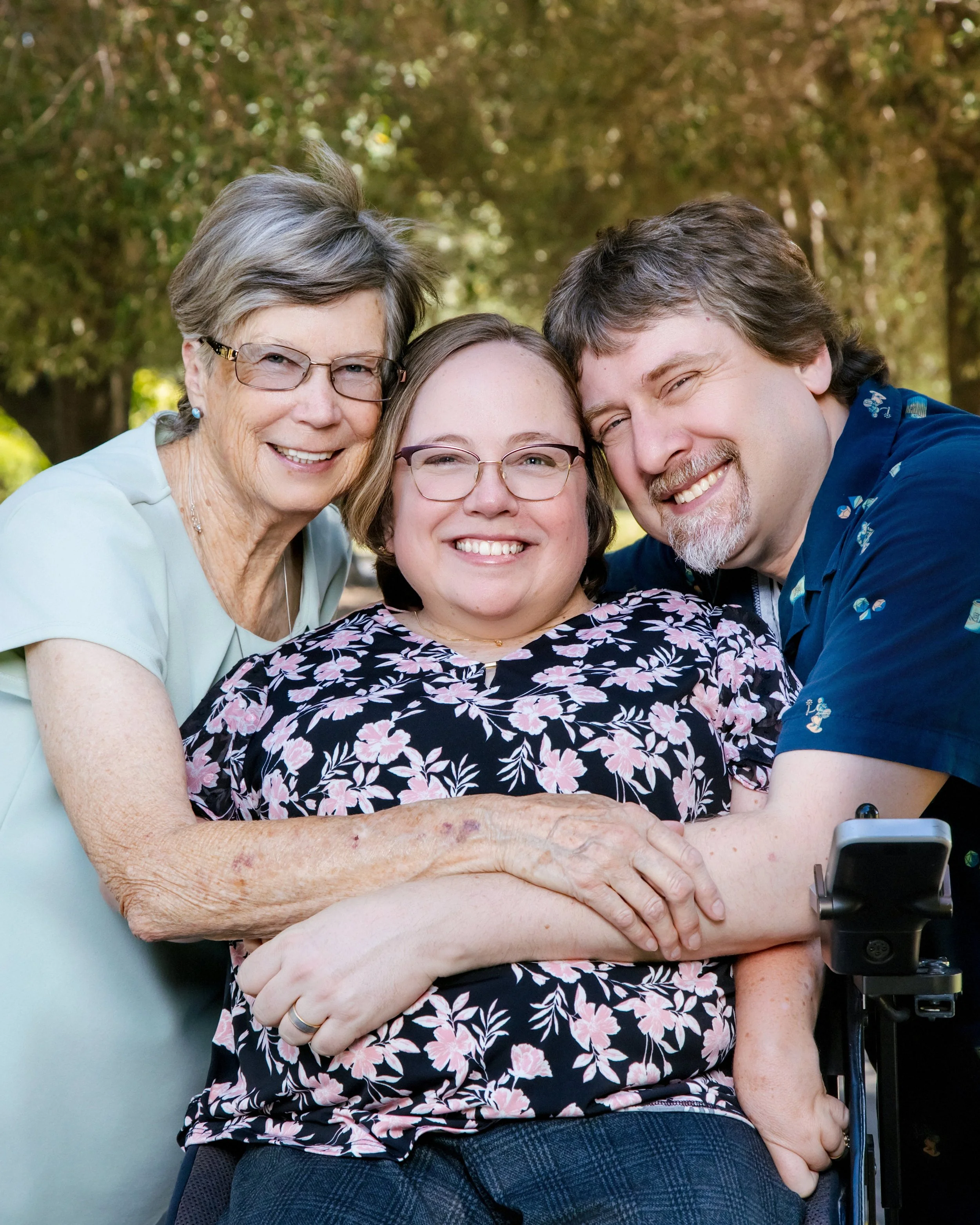 Three people, an elderly woman with short gray hair, glasses, and earrings, a middle-aged woman with glasses and a floral blouse, and a man with gray hair and a beard, smiling and hugging each other outdoors with trees in the background.