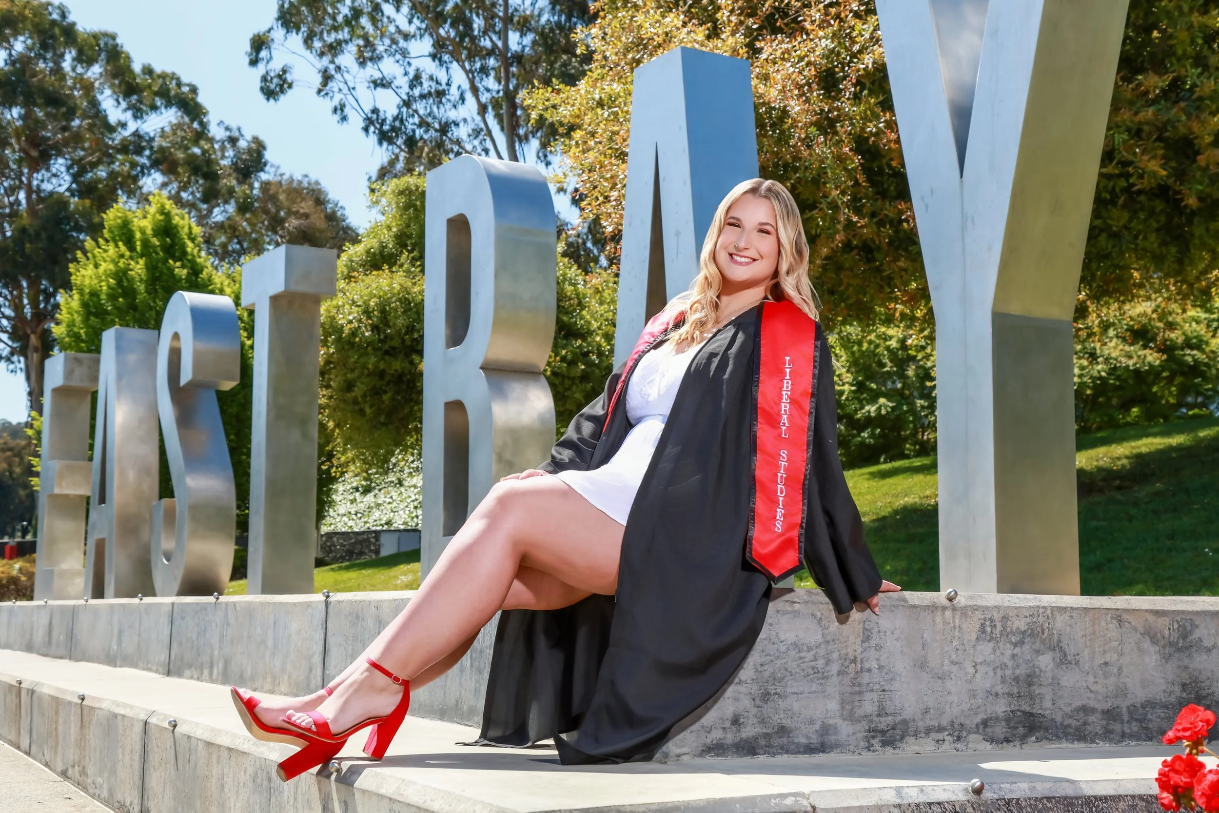 A young woman in a graduation gown and sash sitting on a stone ledge in front of a metallic "UNIVERSITY" sign, smiling and winking at the camera, with trees and a blue sky in the background.