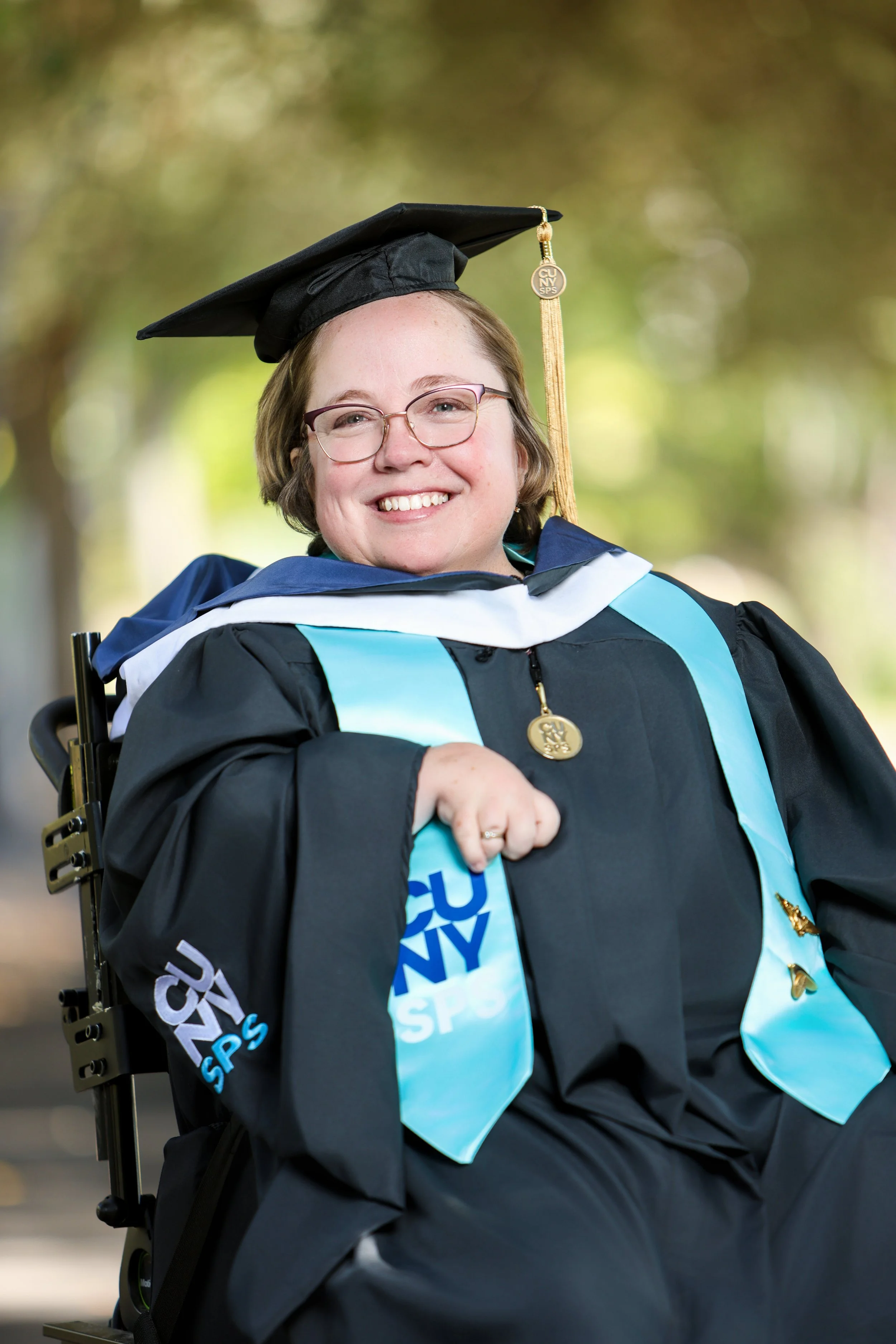 A smiling woman in graduation attire, wearing glasses, a black cap with a gold tassel, and a gown with blue and black accents, seated outdoors with a background of blurred green trees.