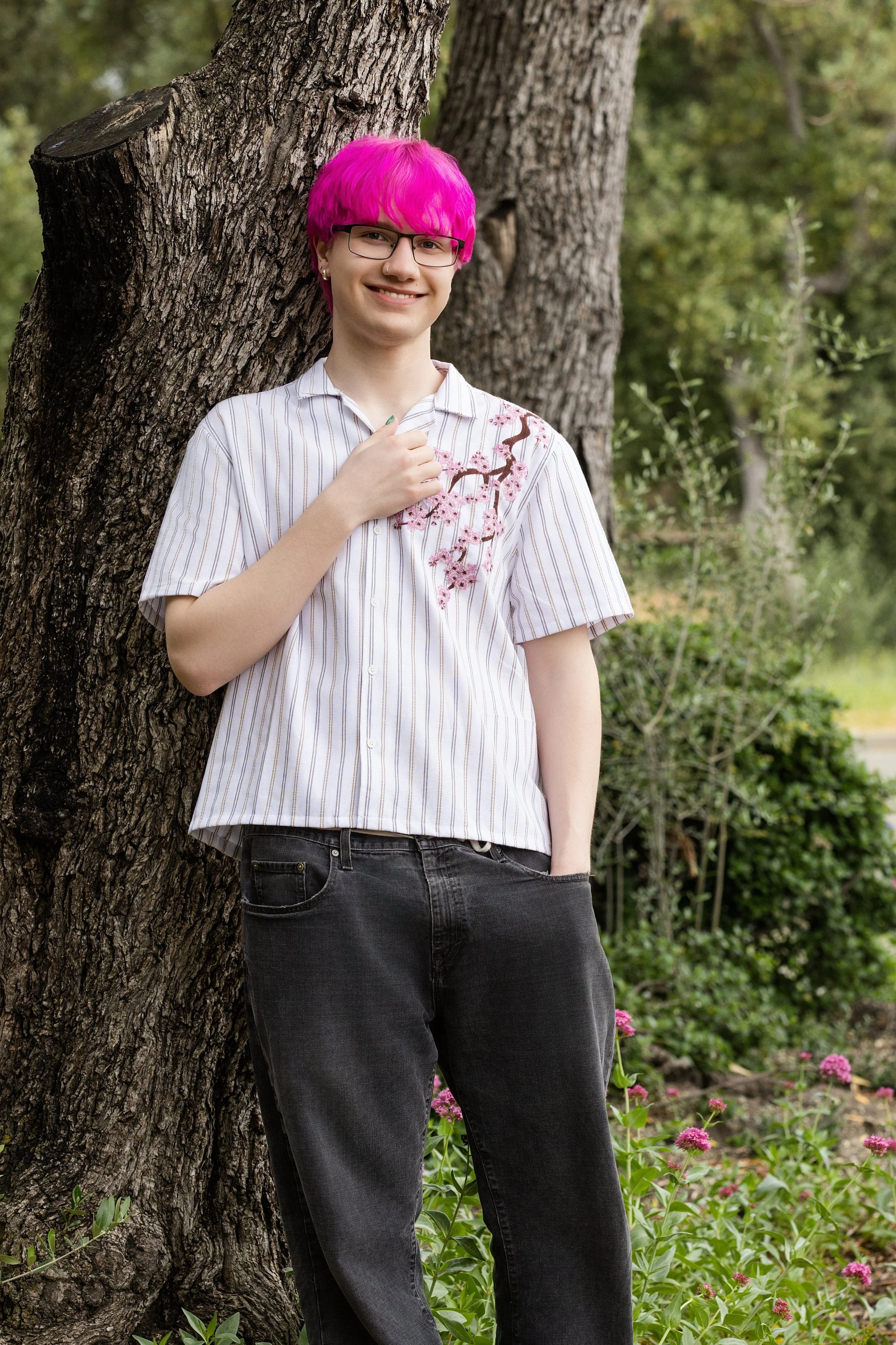 A person with pink hair and glasses leaning against a tree in a garden with pink flowers.