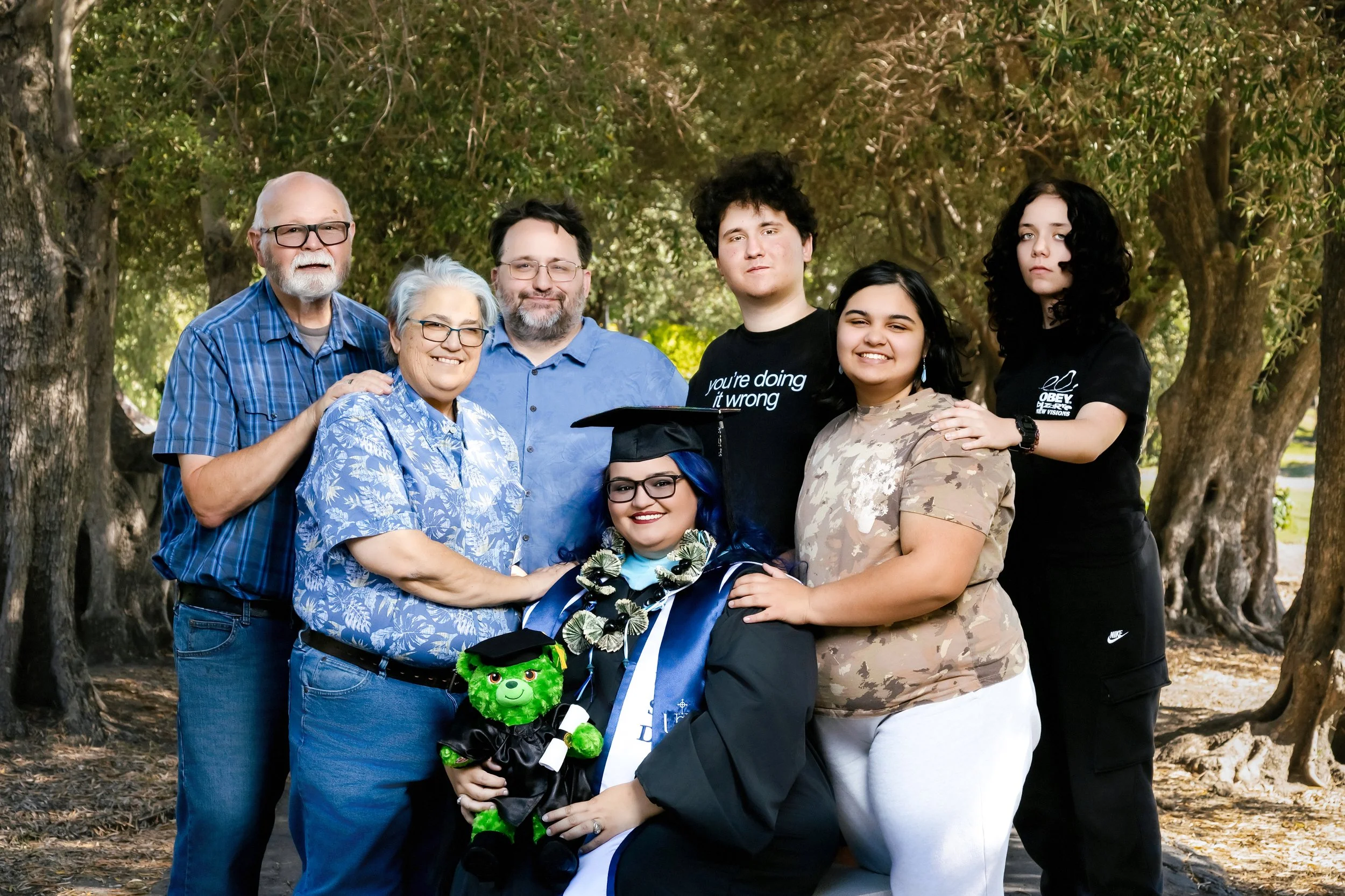 Group of seven people posing outdoors, including a woman in a graduation cap and gown, surrounded by trees.