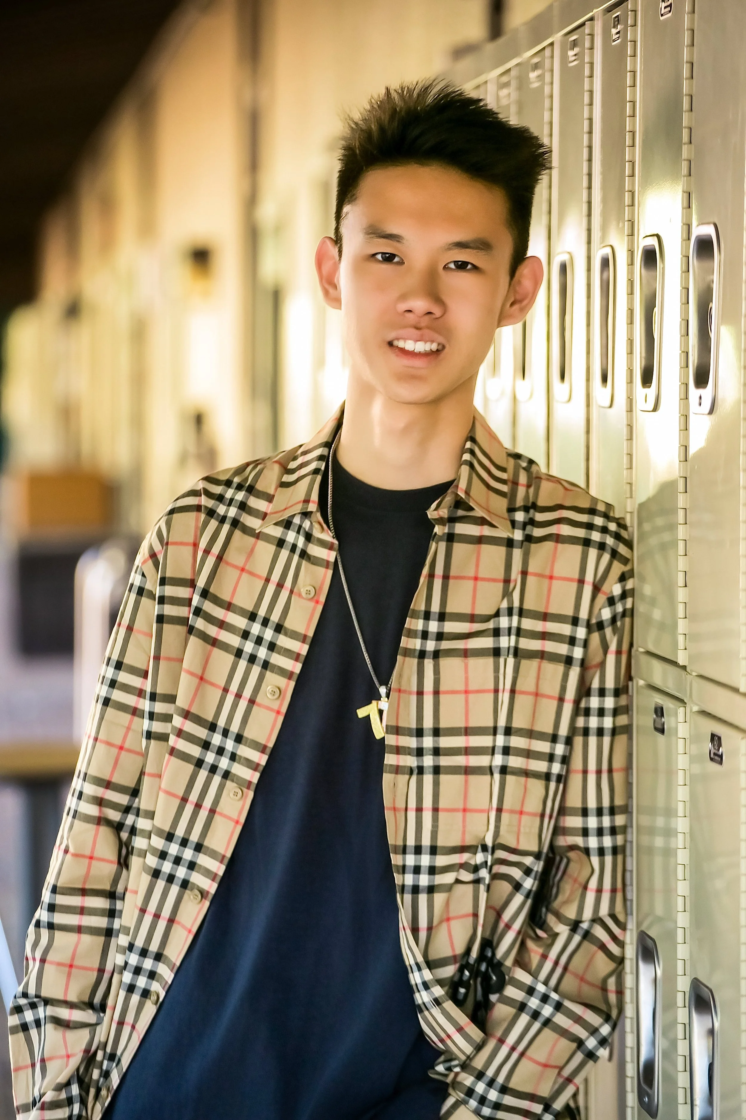 A young man with short black hair smiling and leaning against a row of lockers in a school hallway.
