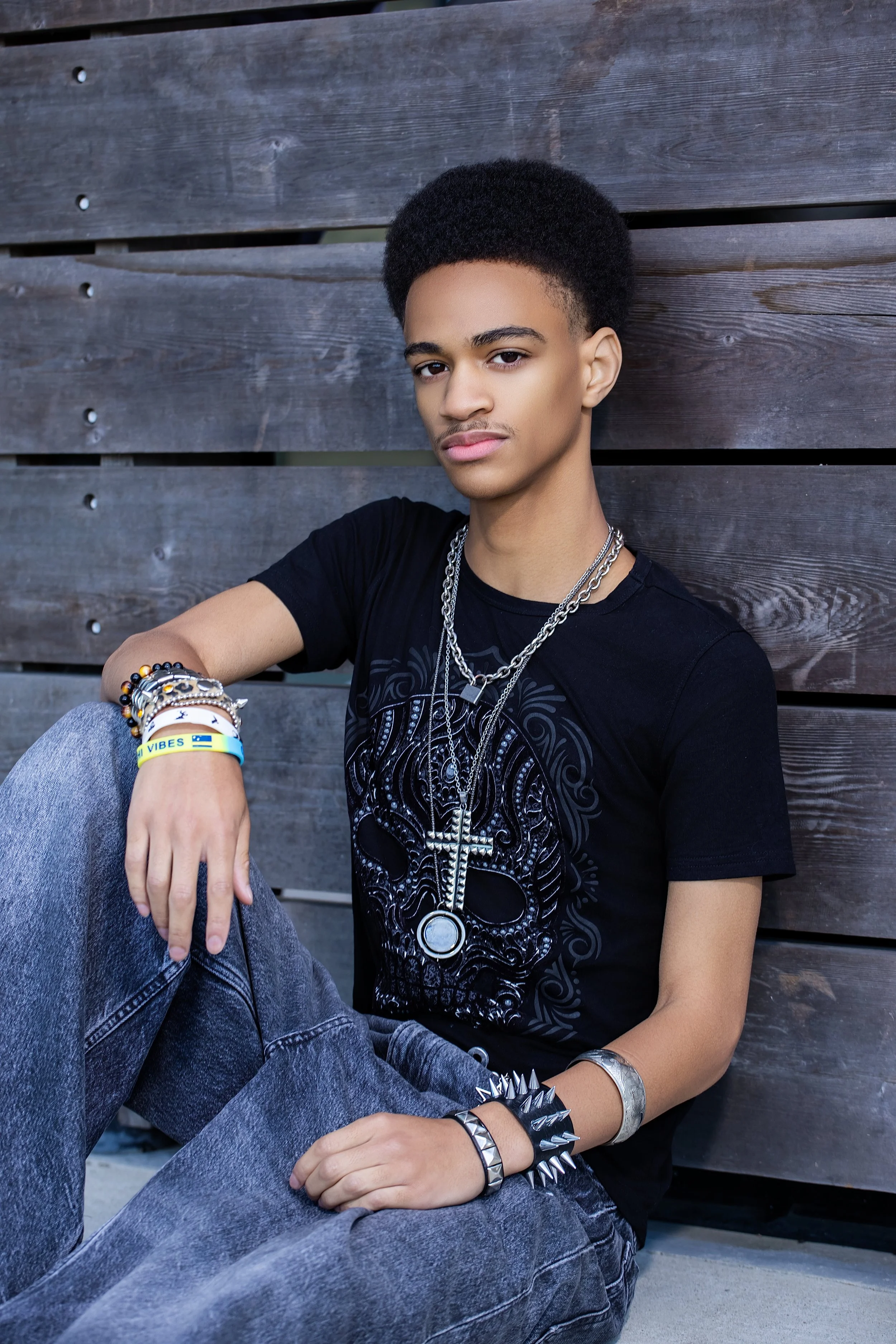 A young man with short curly hair and light brown skin sitting against a wooden fence, wearing a black graphic skull T-shirt, multiple chains, bracelets, and rings, with a relaxed expression.