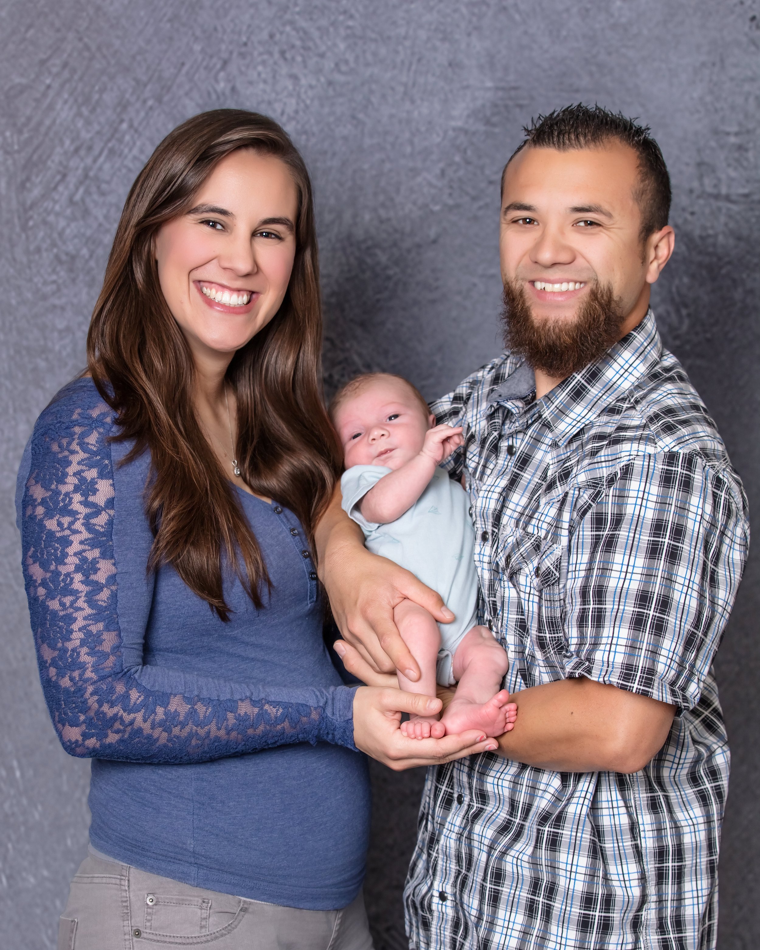 A smiling woman with long brown hair wearing a blue lace top, a smiling man with short spiked hair and a beard holding a baby. The baby has light hair and is wearing a white outfit, looking at the camera.