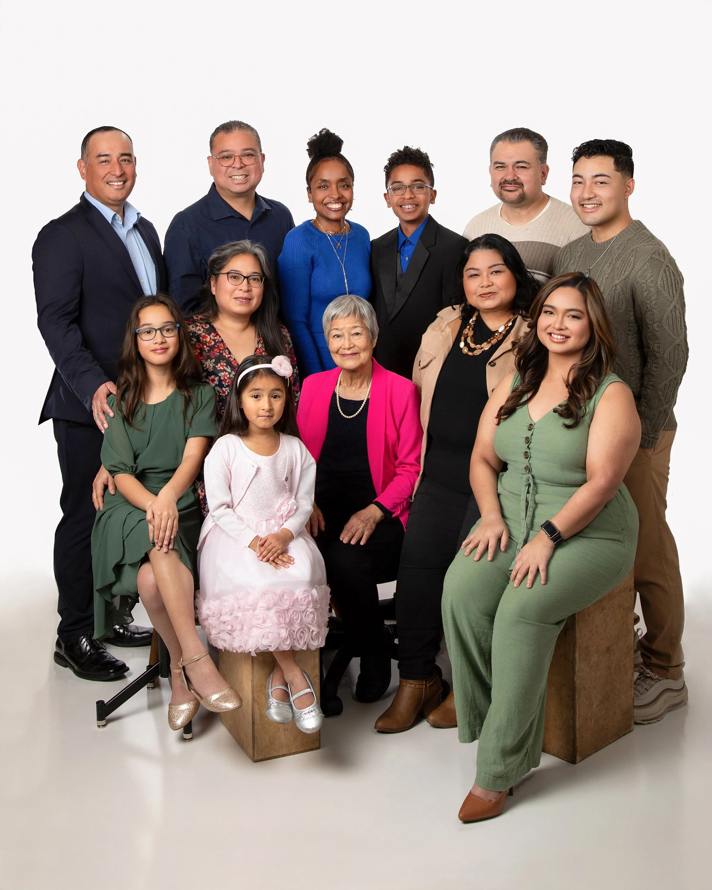 A diverse group of fifteen people of various ages and ethnicities posing together against a plain white background, smiling at camera, dressed in semi-formal attire.