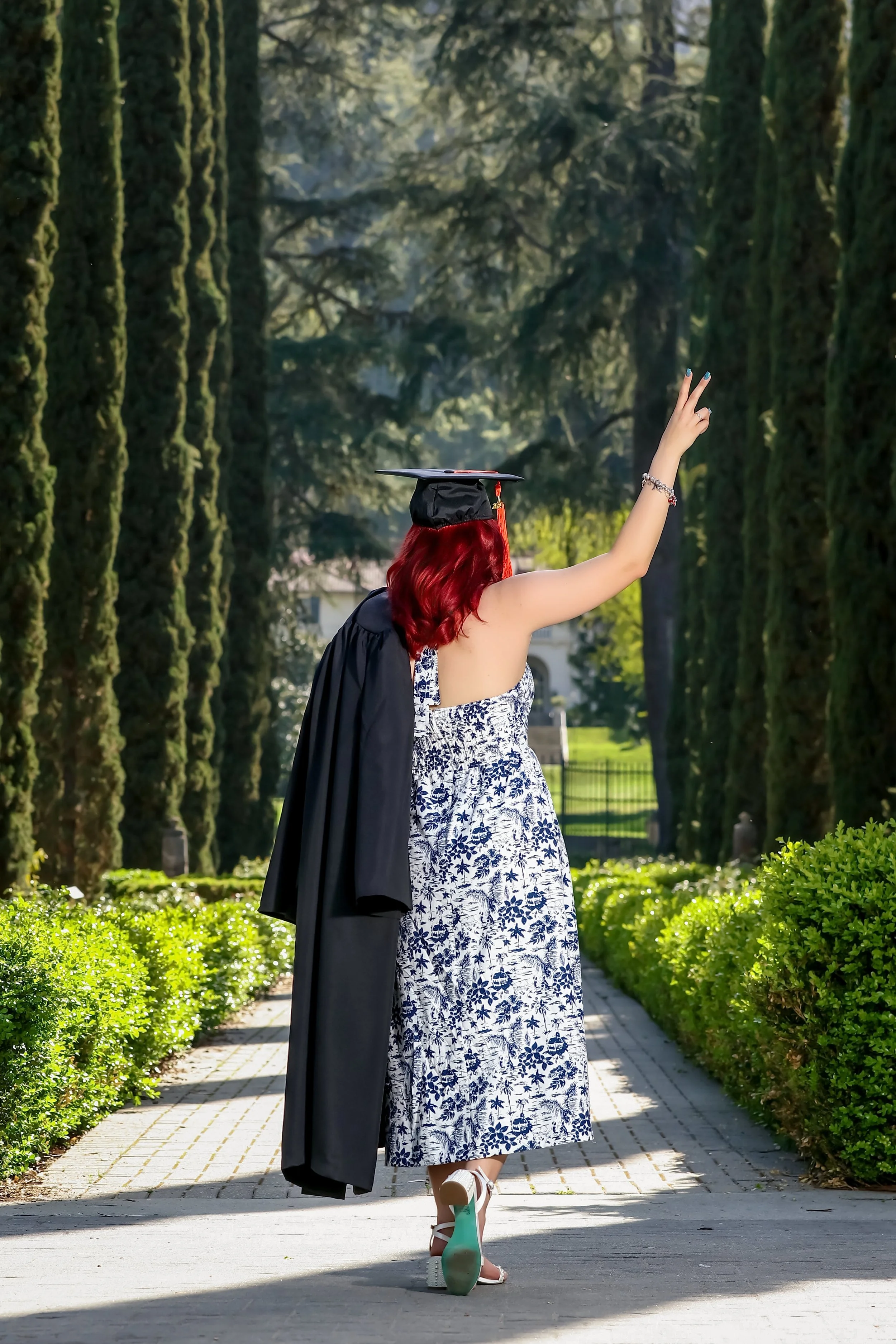 Female college graduate wearing cap and holding gown over the shoulder at an outdoor path surrounded by trees.
