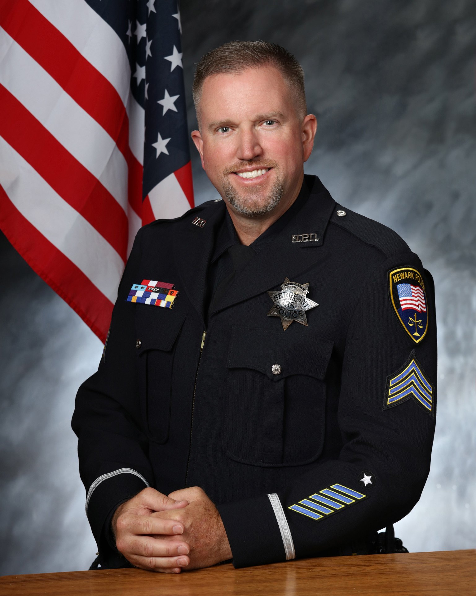 Male police sergeant sitting at desk with American flag and gray studio background.