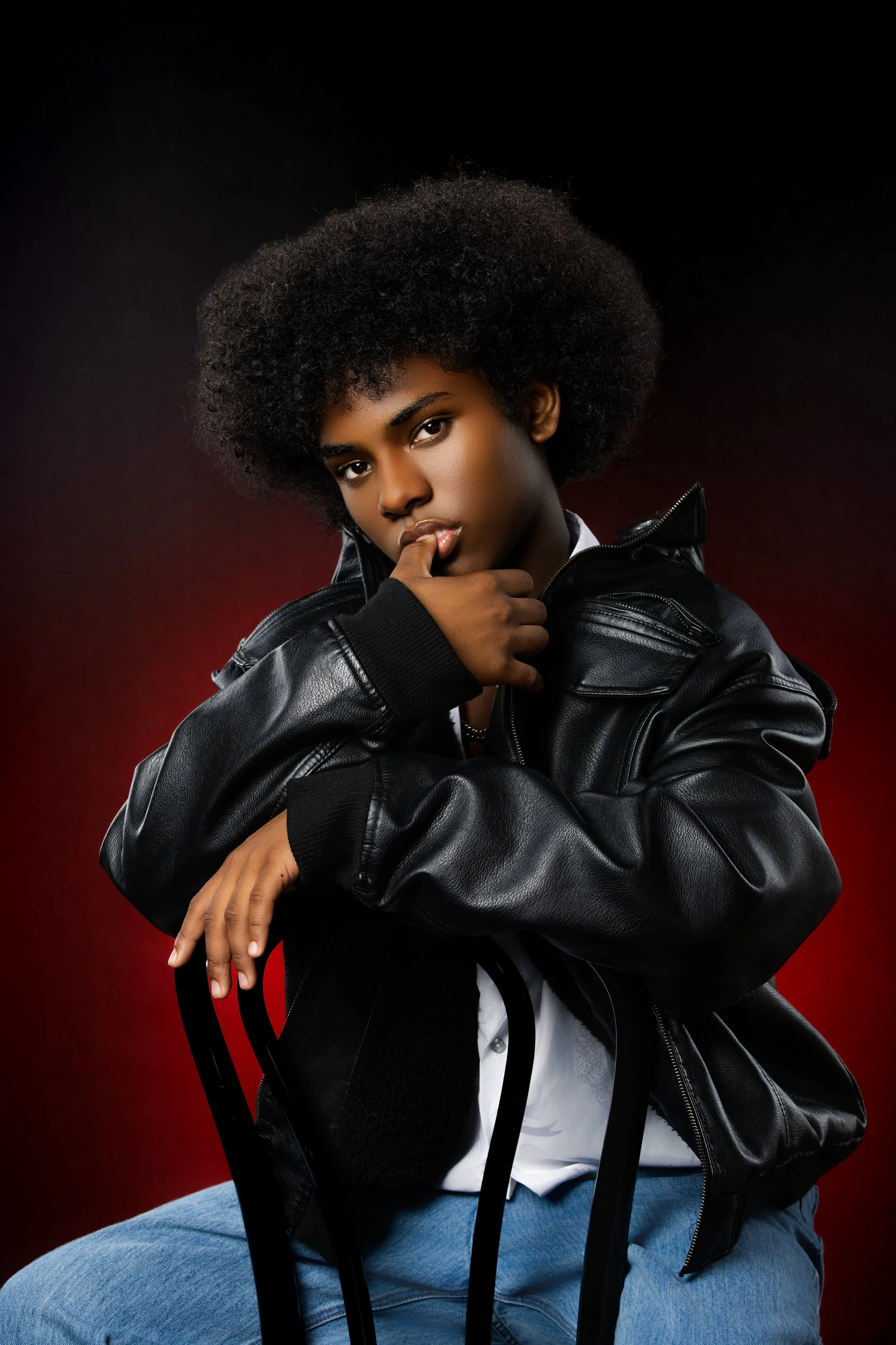 Portrait of a young woman with curly black hair, wearing a black leather jacket, white shirt, and blue jeans, sitting on a black chair against a dark red and black gradient background.