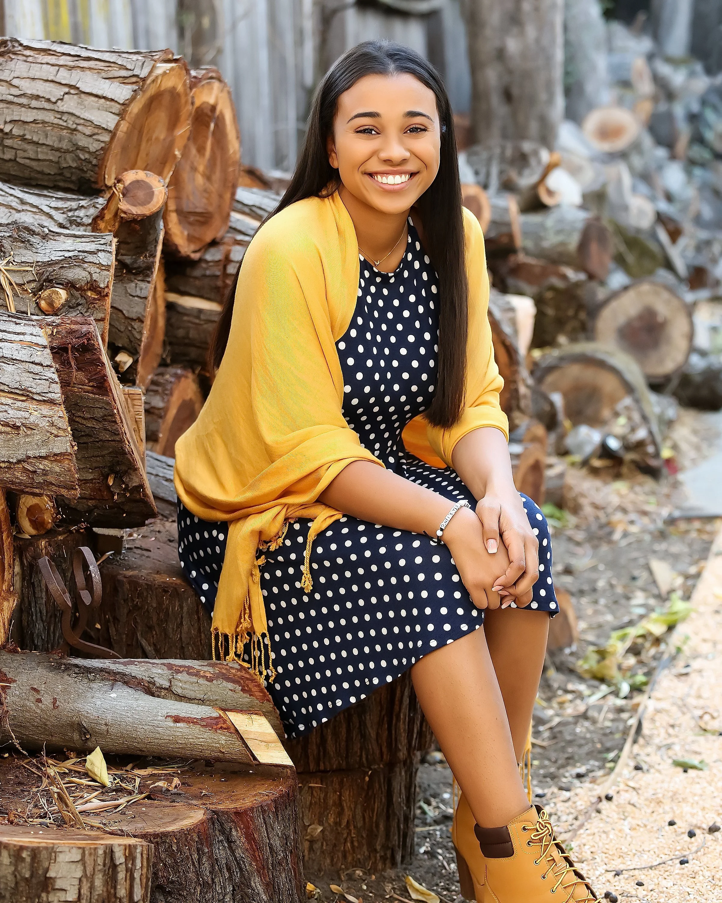 A young woman with long dark hair smiling and sitting on a large tree stump in a wooded outdoor area. She is wearing a navy blue polka dot dress, a yellow cardigan, and yellow hiking boots, with a stack of chopped firewood behind her.