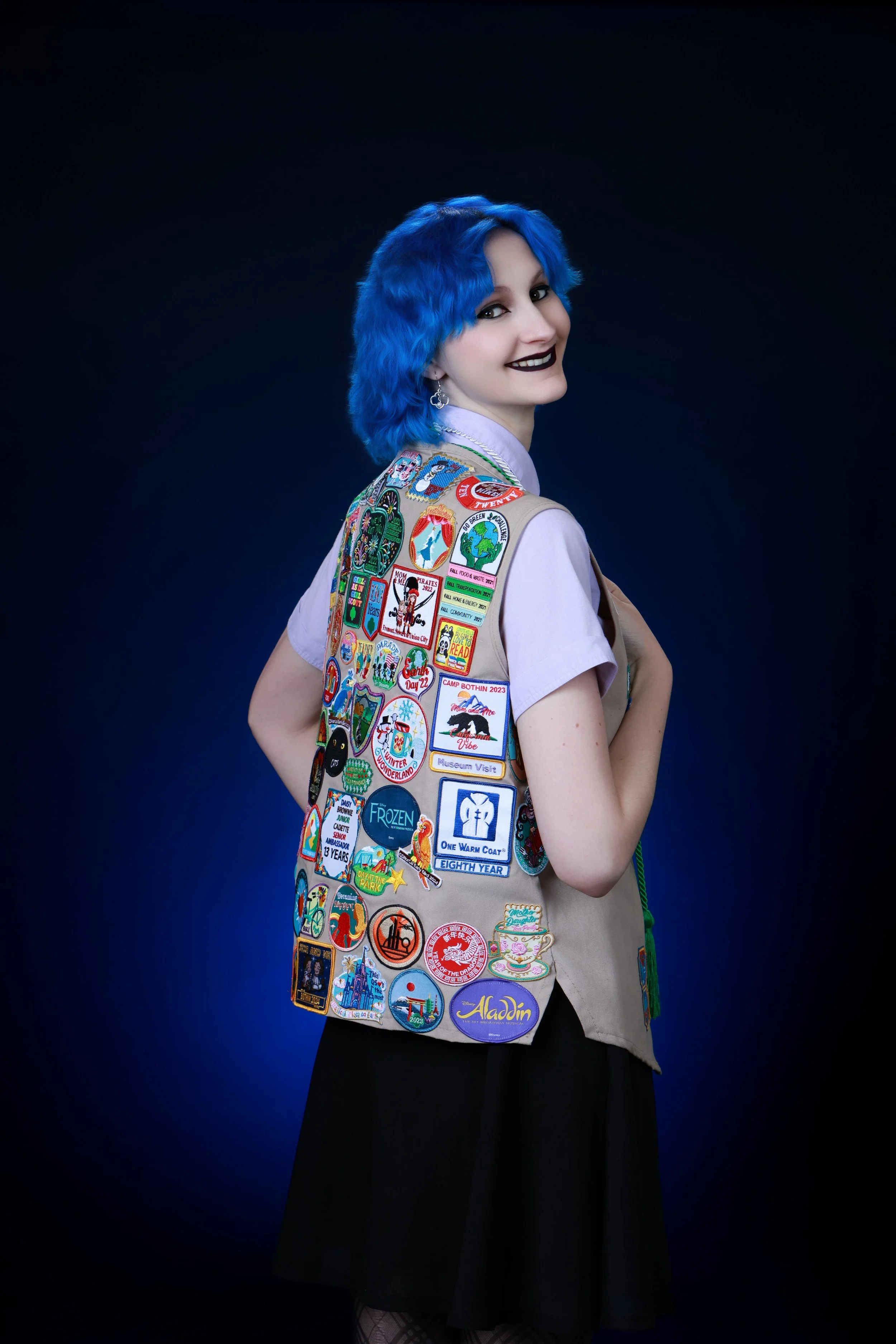 A woman with bright blue hair wearing a tan vest covered in colorful patches, a light purple shirt, and a long black skirt, standing against a dark background.