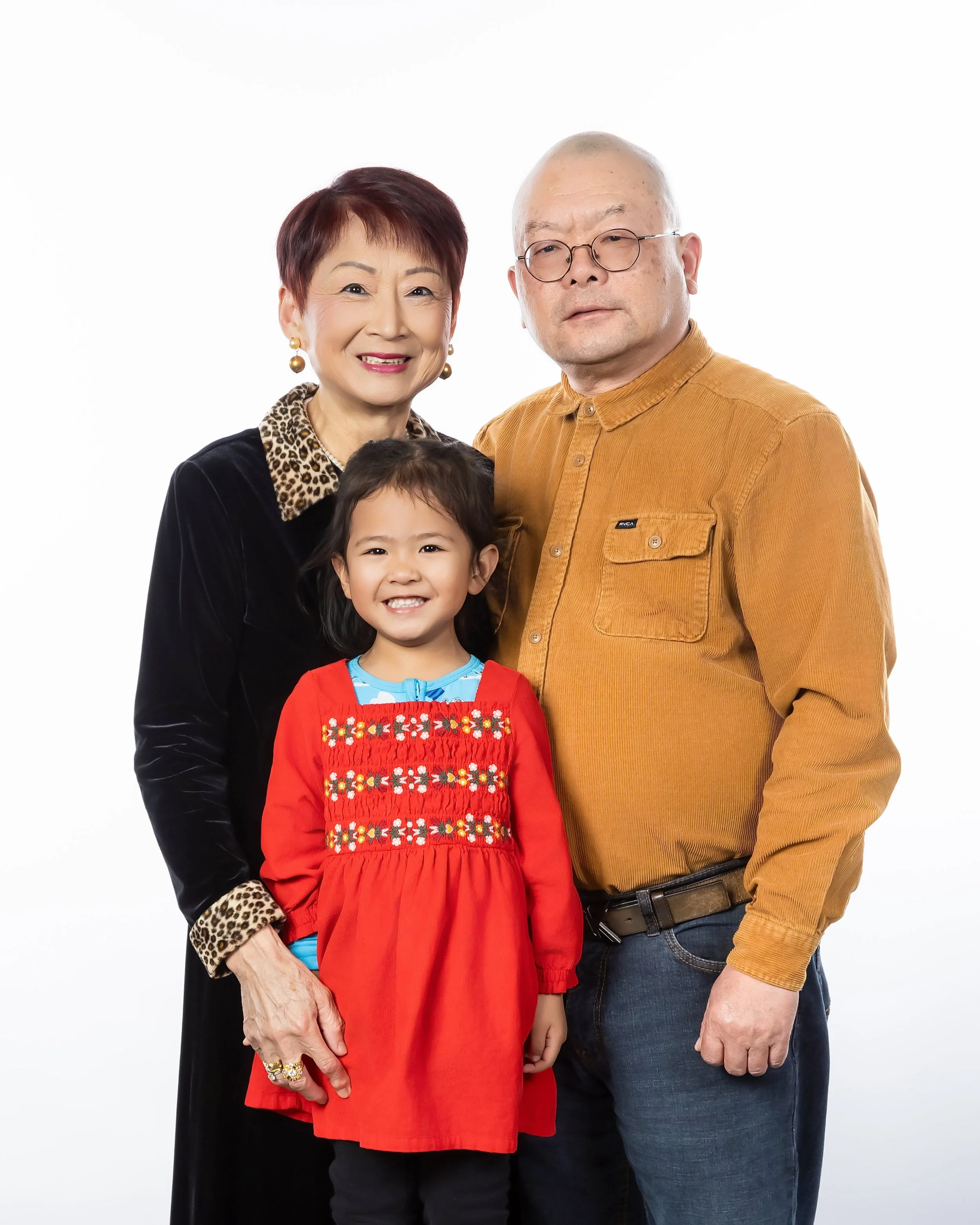 A multigenerational family portrait featuring a smiling older woman with short red hair, jewelry, and a black top with leopard print collar; a middle-aged man with glasses, a beige shirt, and dark jeans; and a young girl with dark hair, wearing a red
