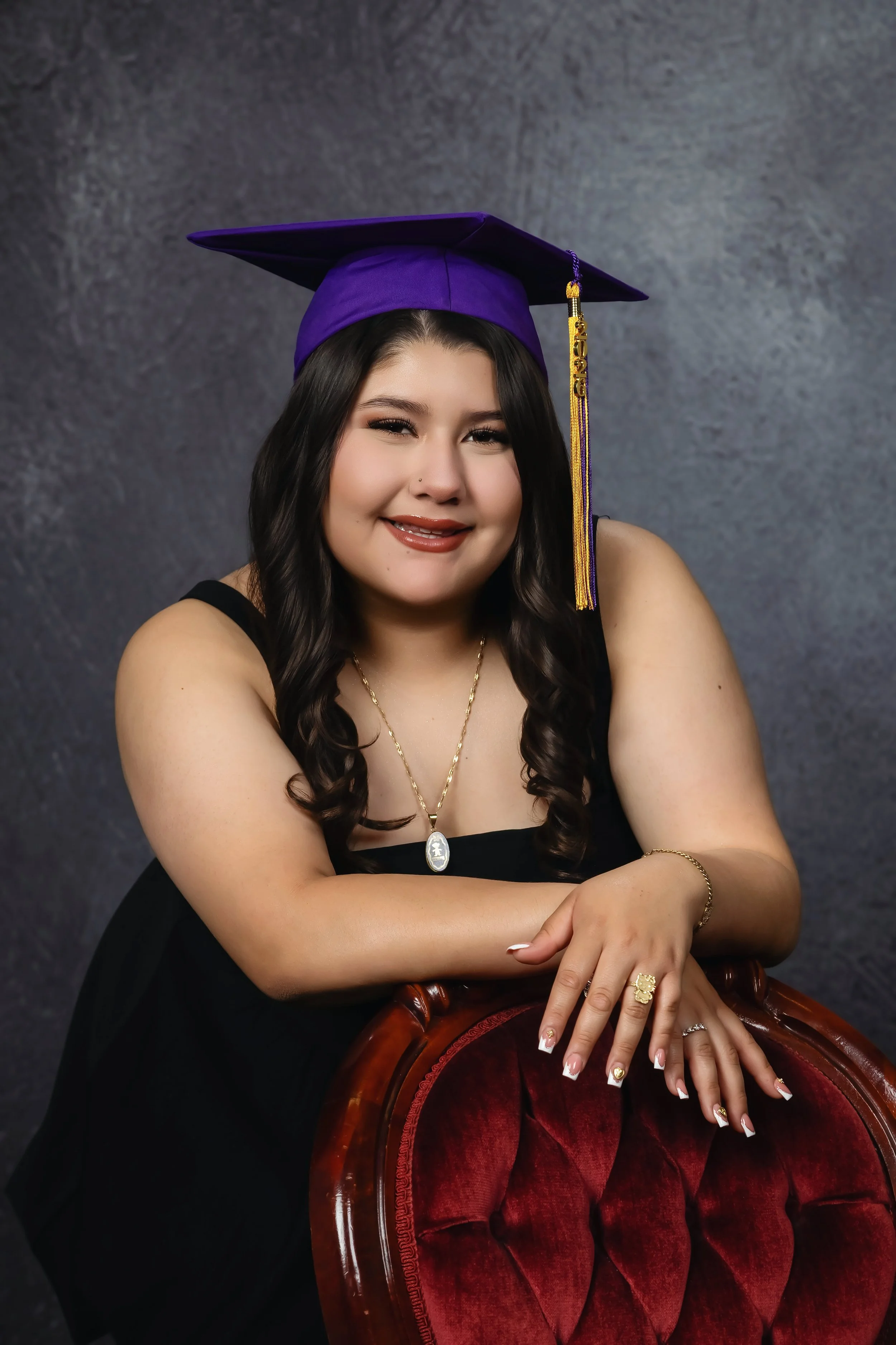 A young woman wearing a purple graduation cap and gown, with a gold tassel, sitting indoors against a gray wall. She has long dark hair, makeup, and jewelry, and is smiling with her arms crossed, resting her hands on the back of a wooden chair with r