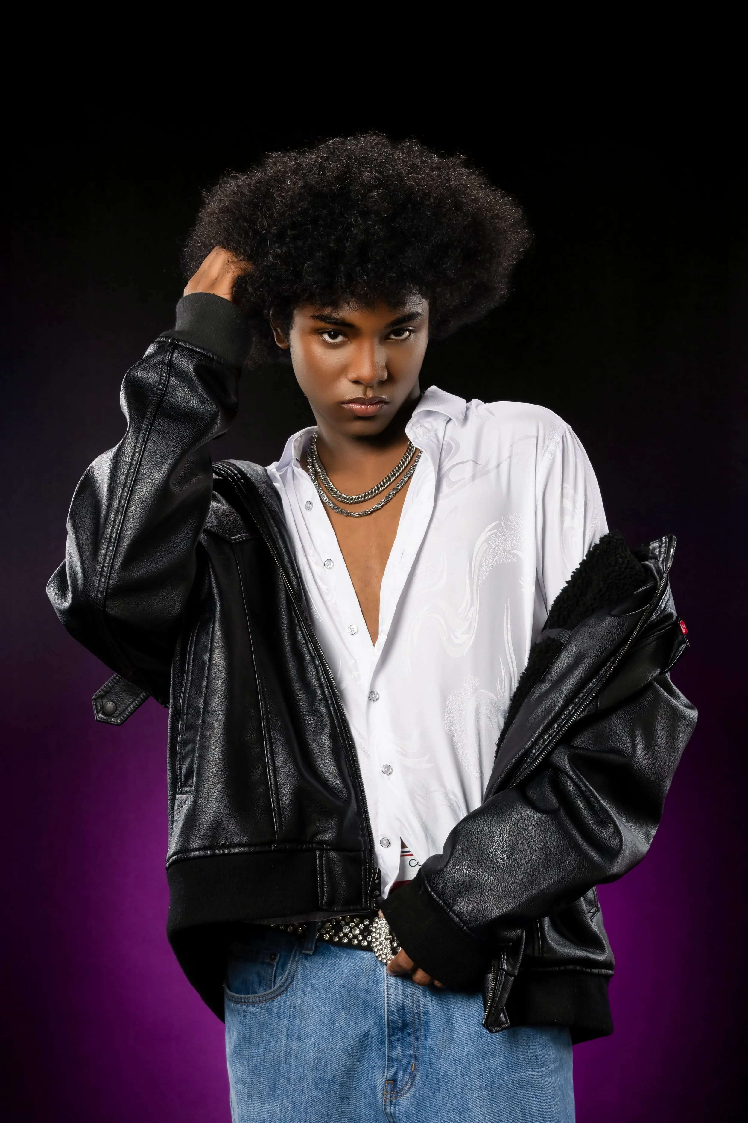 Young woman with dark skin and an afro hairstyle wearing a black leather jacket, white shirt, layered necklaces, and blue jeans, standing against a dark background with purple lighting at the bottom.