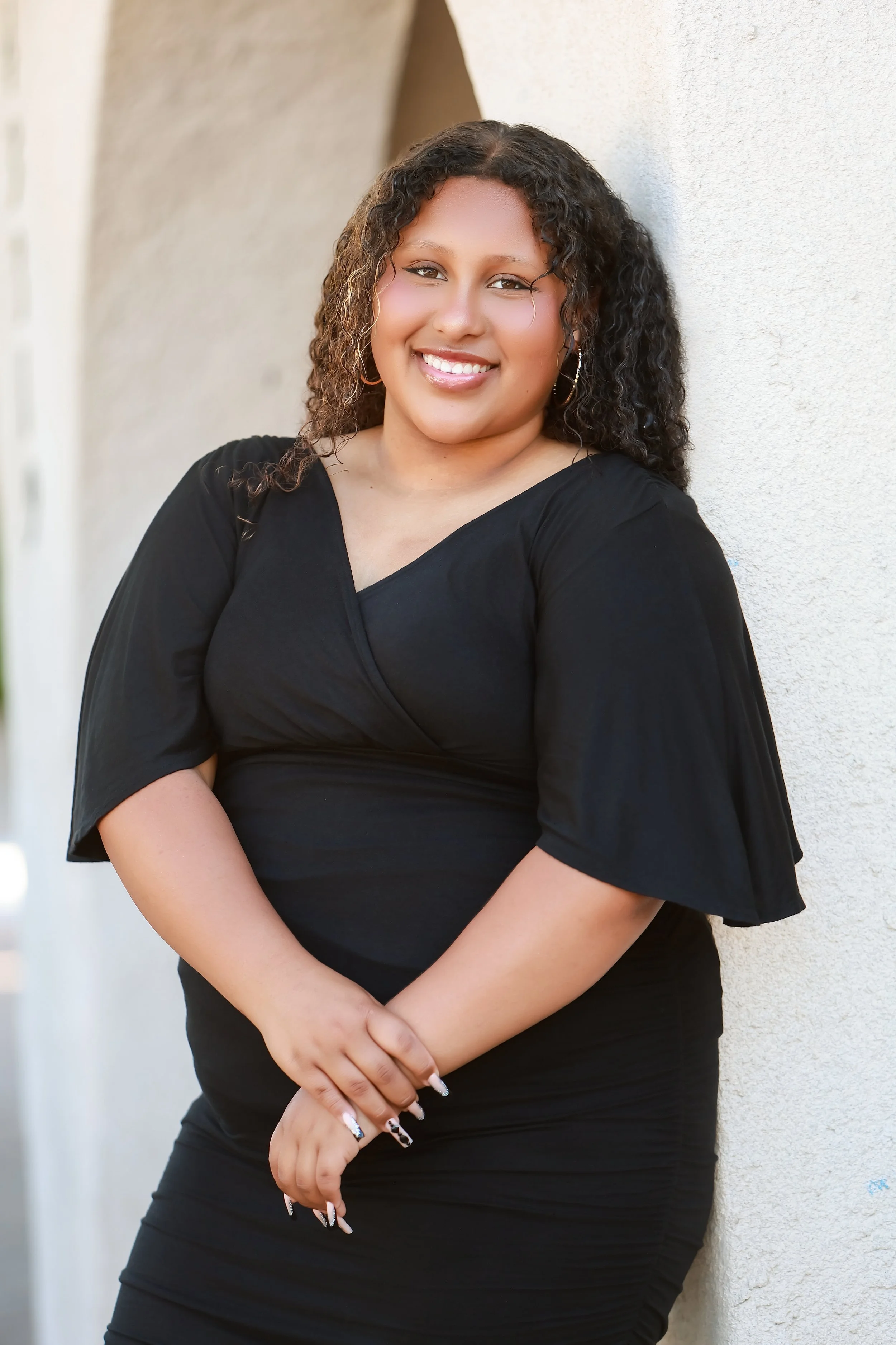 A woman with curly hair wearing a black dress, smiling and leaning against a beige wall.