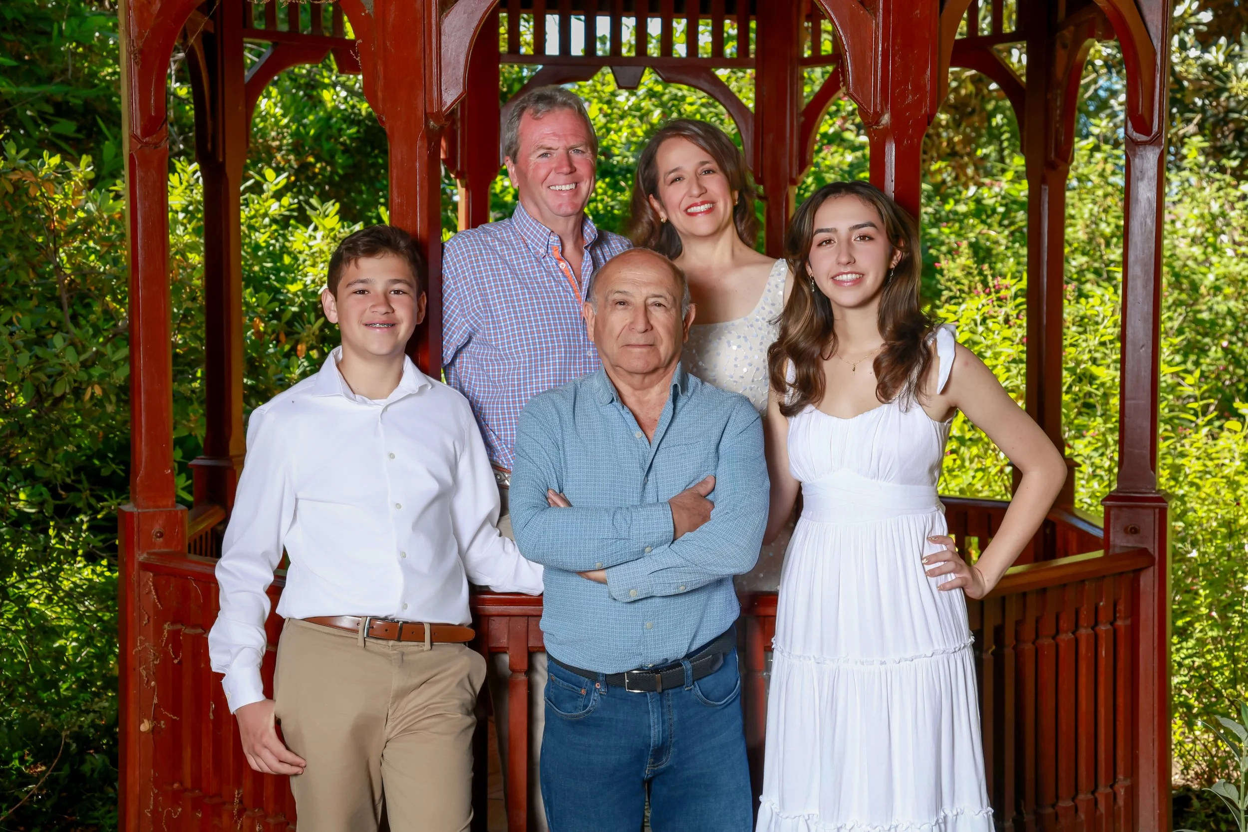 A multigenerational family of six people standing on a wooden porch surrounded by greenery. They are smiling and posing for a photo.