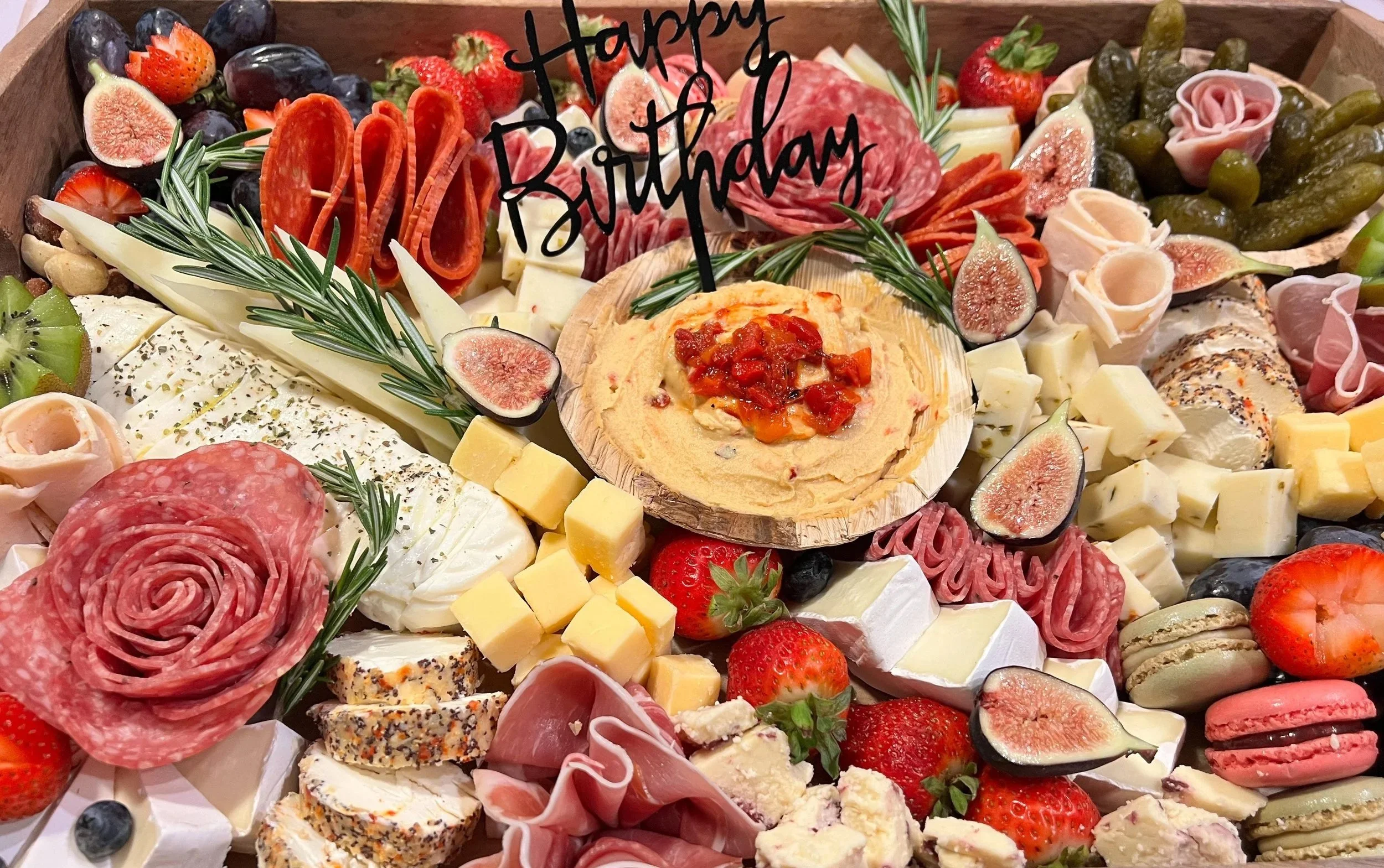 A large wooden platter of assorted cheeses, cured meats, fresh berries, fruits, pickles, and crackers, with a small wooden bowl of hummus topped with chopped tomatoes at the center, and a handwritten “Happy Birthday” sign.