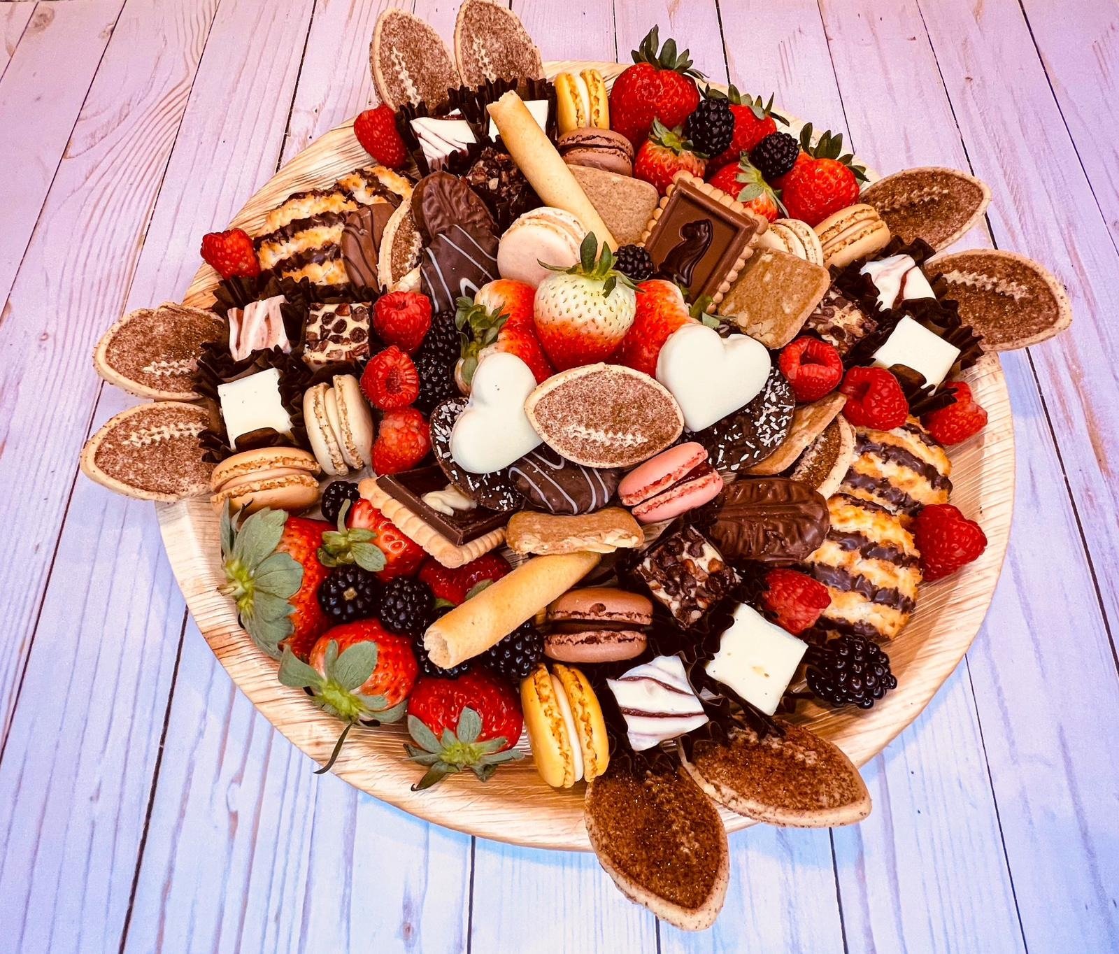 A large round wooden platter filled with assorted chocolates, strawberries, raspberries, blackberries, macarons, and cookies on a purple wooden surface.