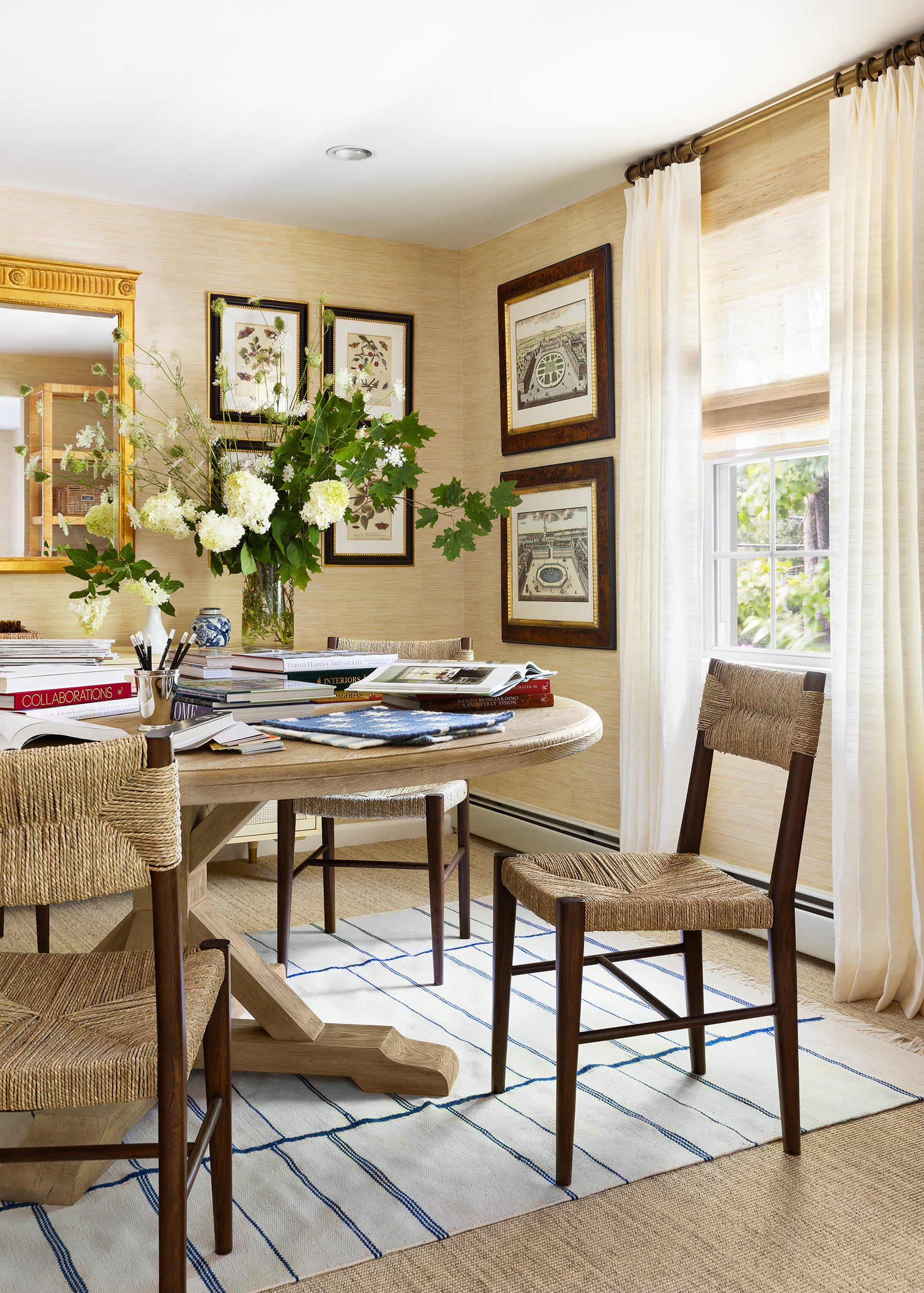 A dining room with a wooden table, four chairs with woven seats, an open magazine, and a white rug with blue lines. There is a large flower arrangement in a glass vase, and framed artwork on beige walls. A window with white curtains lets in natural light.