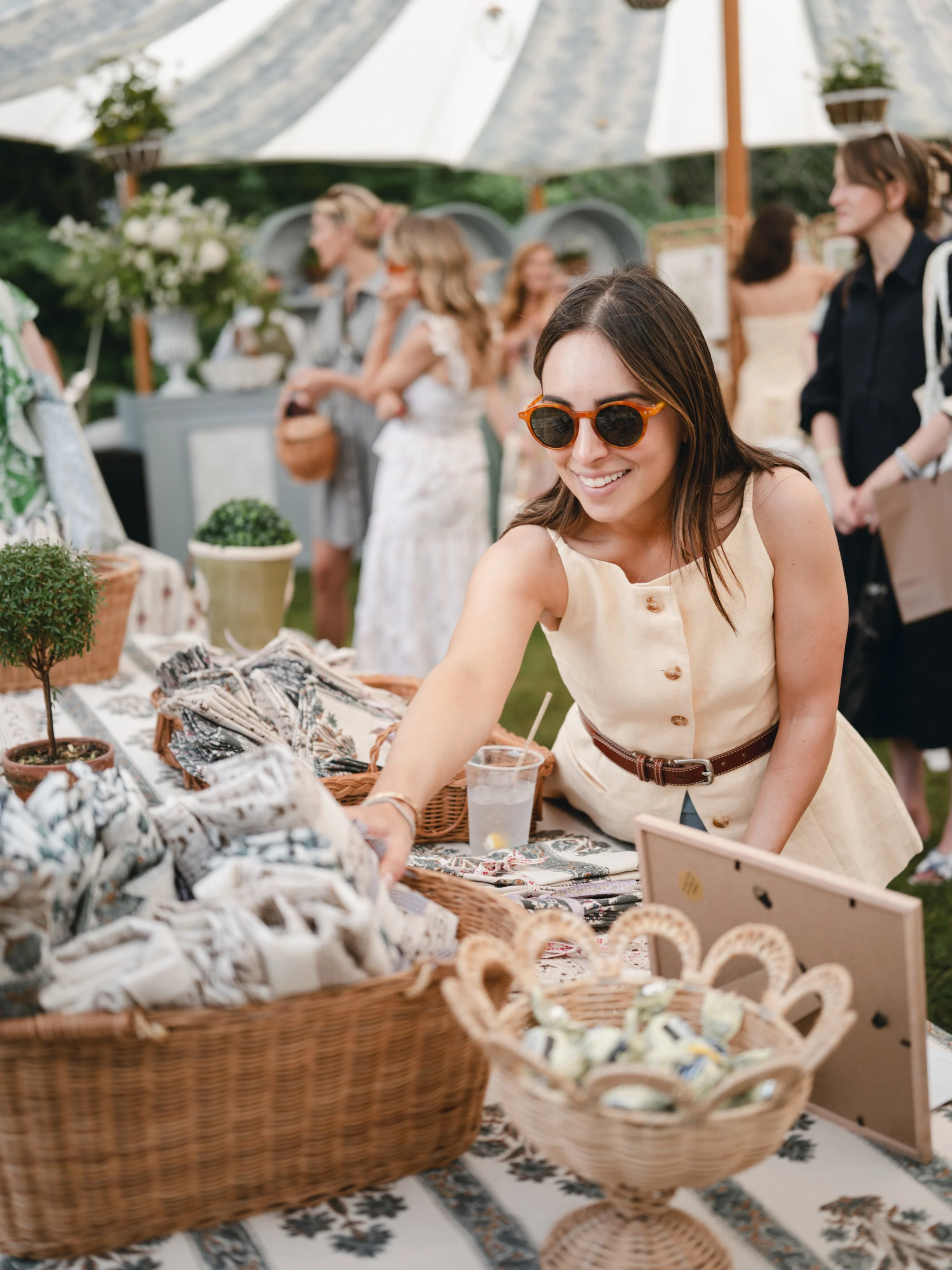 A woman with brown hair and orange sunglasses smiling at a market stall displaying various textiles, jewelry, and crafts under a large white patio umbrella.