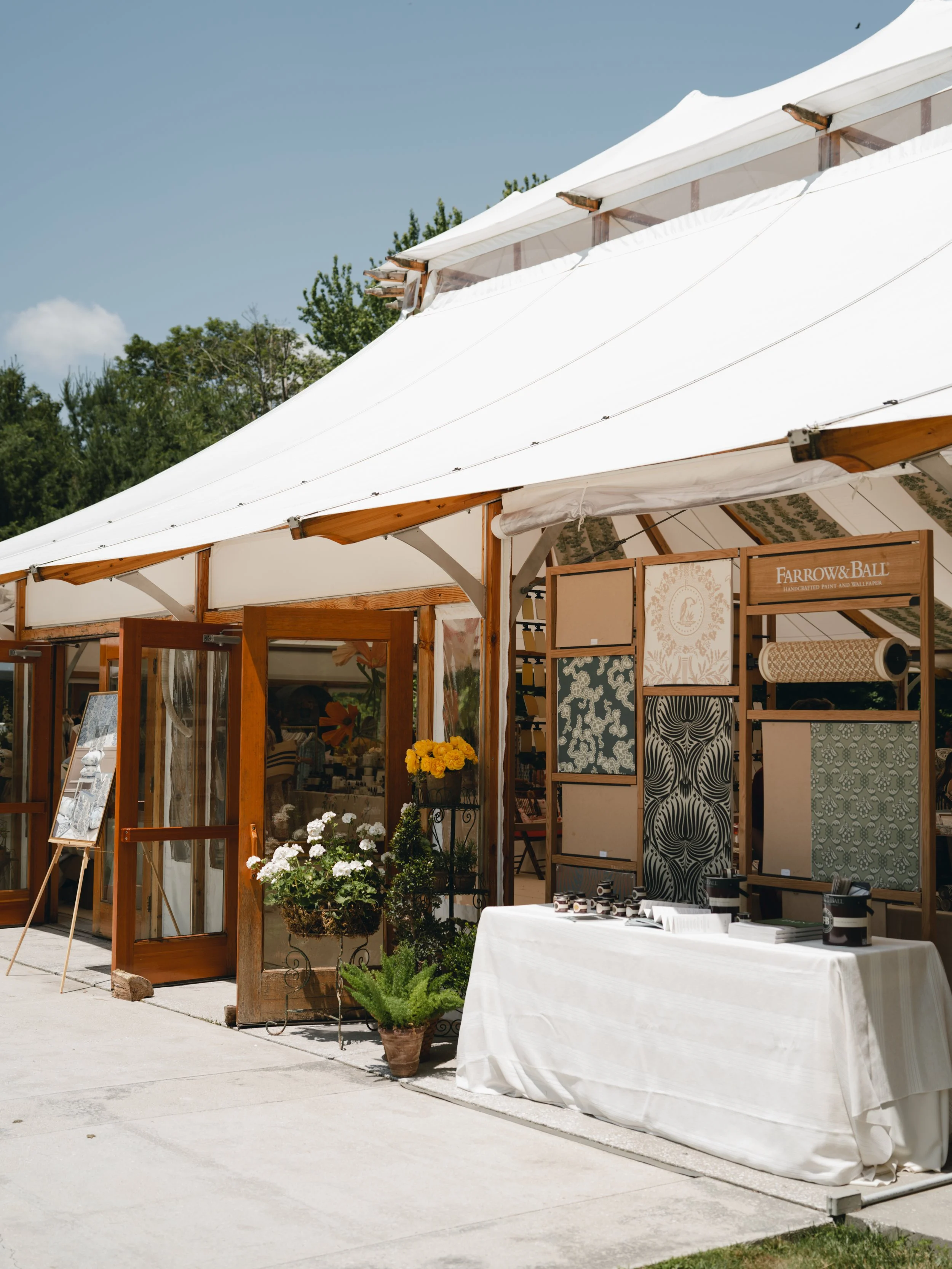 Outdoor market or craft fair booth with wooden frame, white canopy, patterned wallpaper displays, plants, and table with sale items.