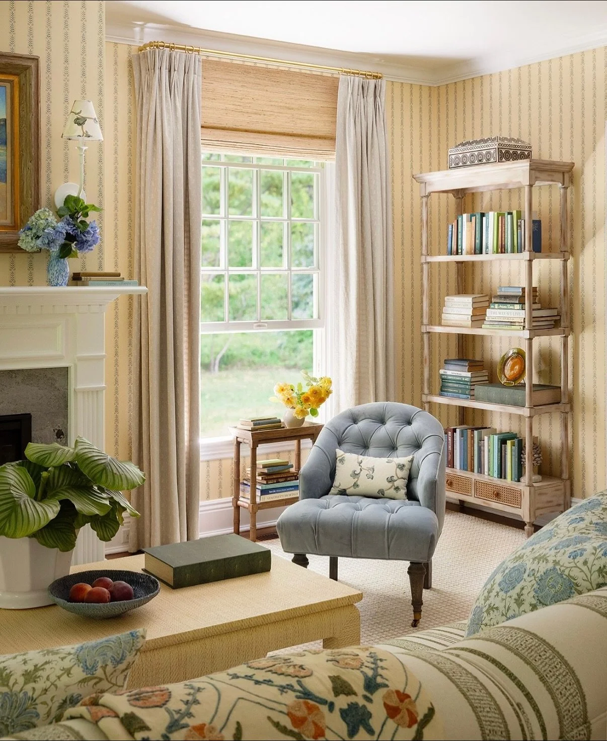 A cozy living room with a large window, beige curtains, a light blue tufted armchair with a decorative pillow, a wooden side table with books and yellow flowers, a white fireplace with a vase of blue and green hydrangeas, a tall bookshelf filled with books and decorative items, and a coffee table with a bowl of peaches and a closed book.