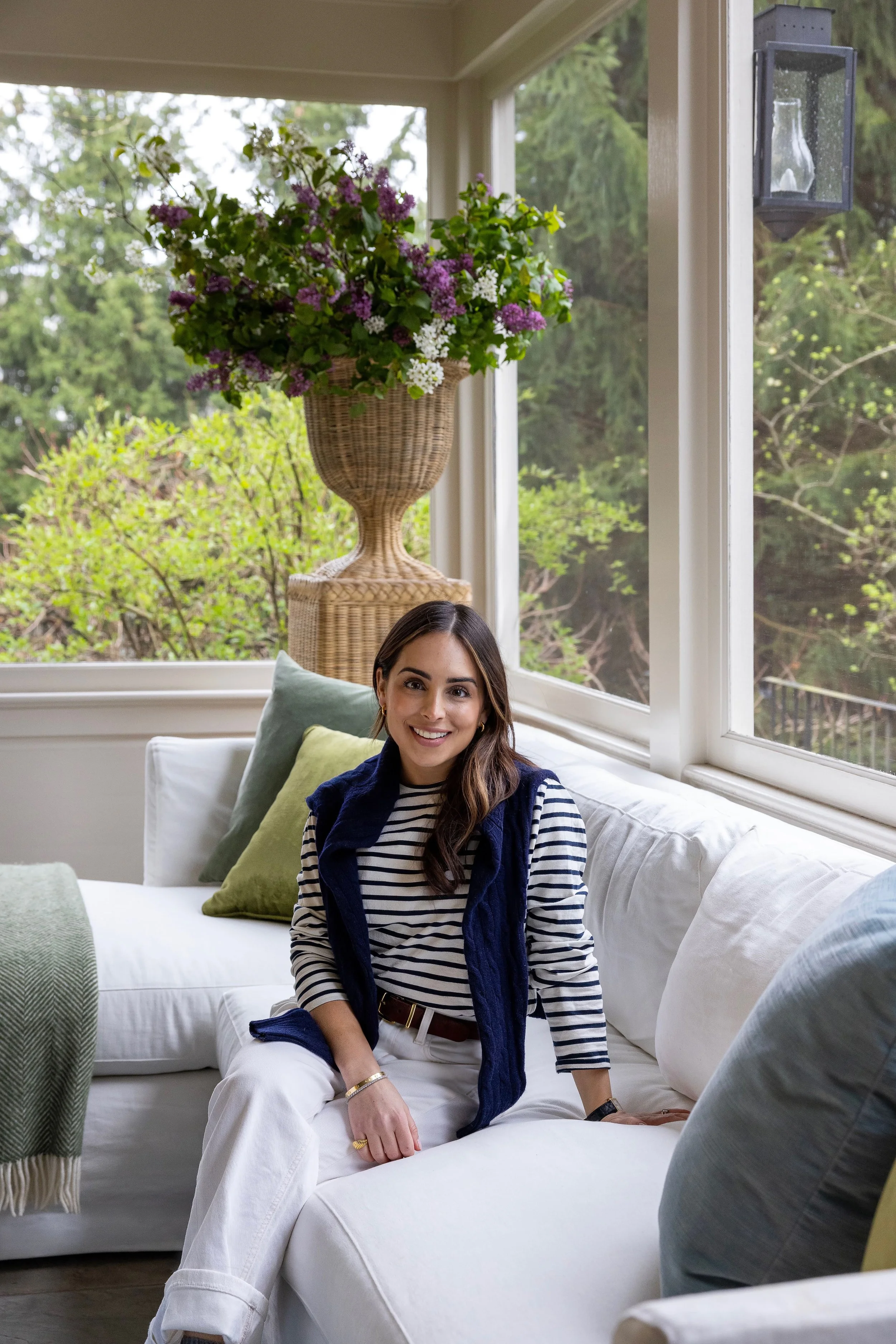 A woman with long brown hair, wearing a striped shirt and white pants, sitting on a white sofa in a bright room with large windows and a view of greenery outside. There are cushions on the sofa and a green blanket draped over the armrest.