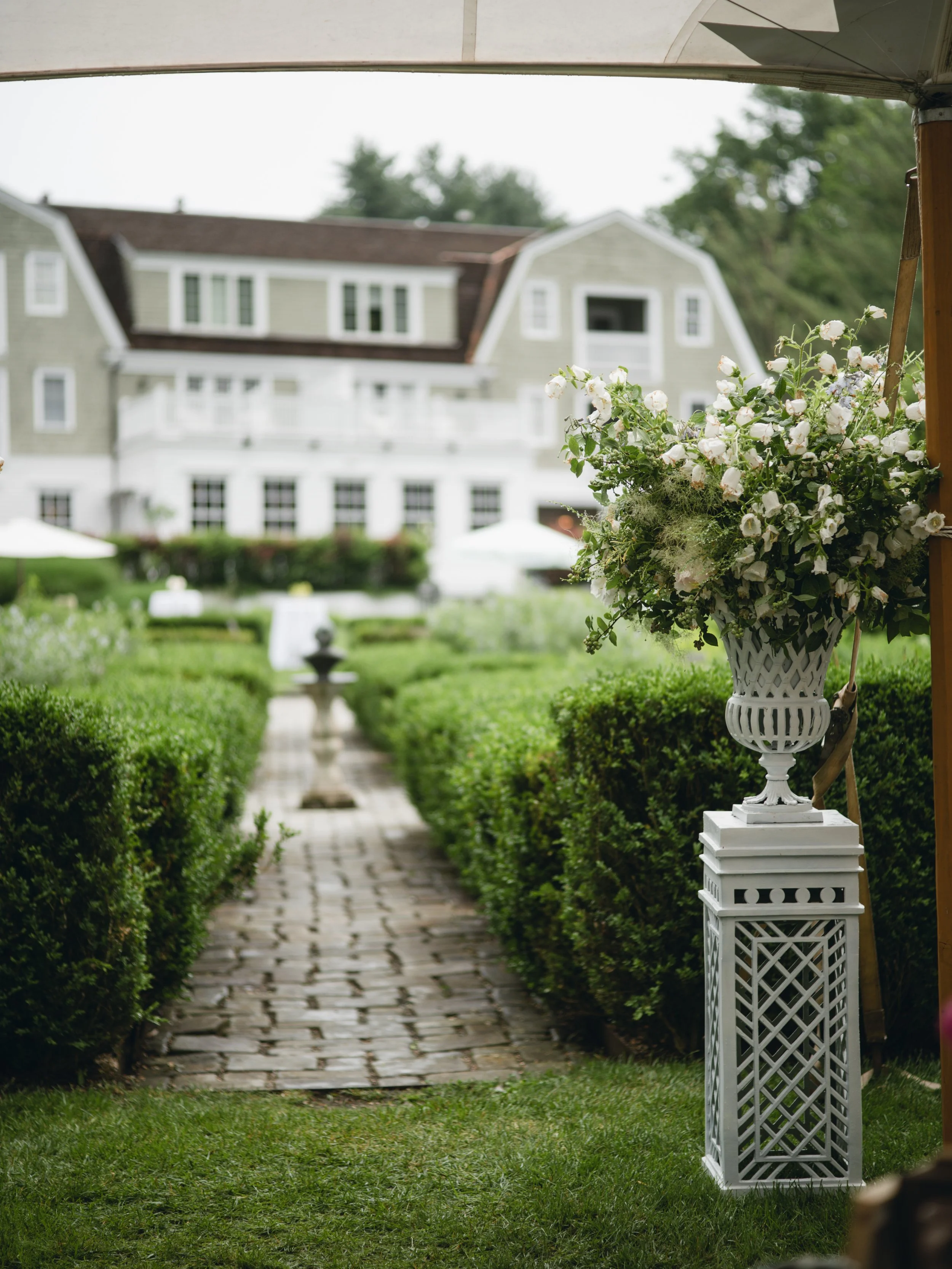 White flower arrangement in a white decorative planter placed on a tall, lattice-style stand, located on a grassy lawn near a stone pathway, with a mansion in the background.