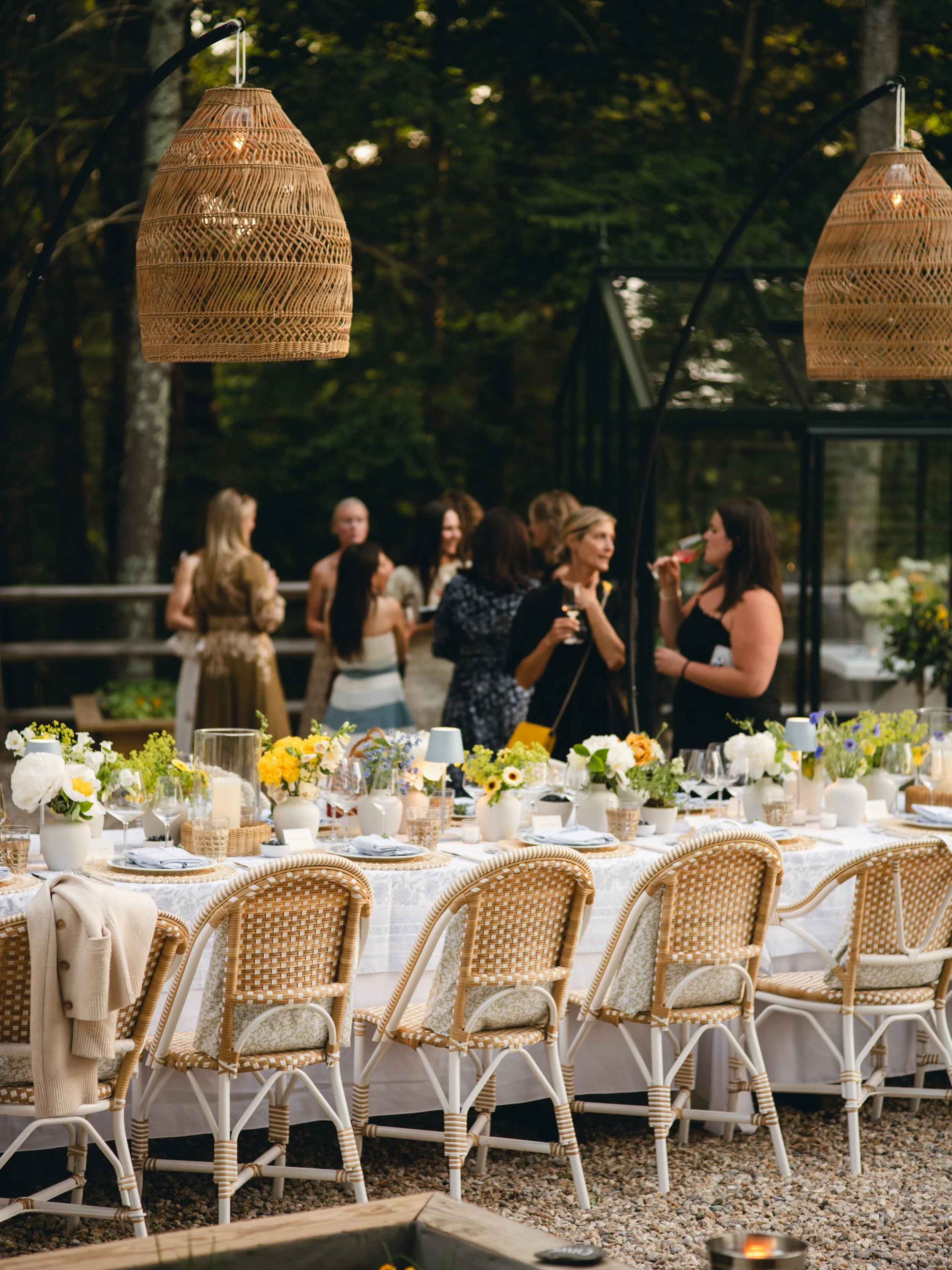 An outdoor dinner setup with a long table decorated with numerous floral arrangements, candles, and tableware. Surrounding chairs are woven in a light rattan pattern. In the background, a group of women are standing and socializing, with trees and a 