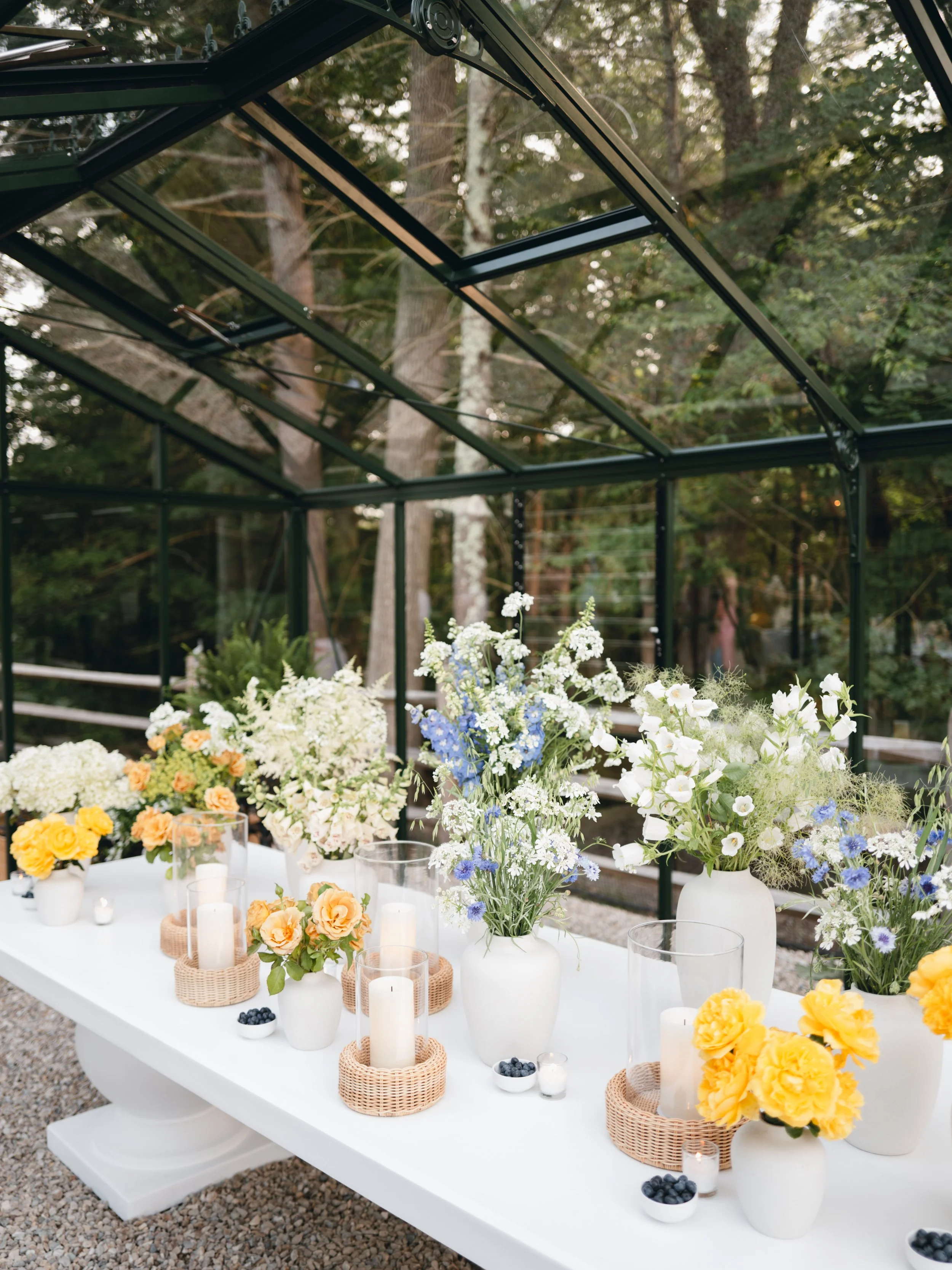 A white table set outdoors with floral arrangements in white vases, candles, and small bowls of blueberries, surrounded by a black metal greenhouse structure and trees in the background.