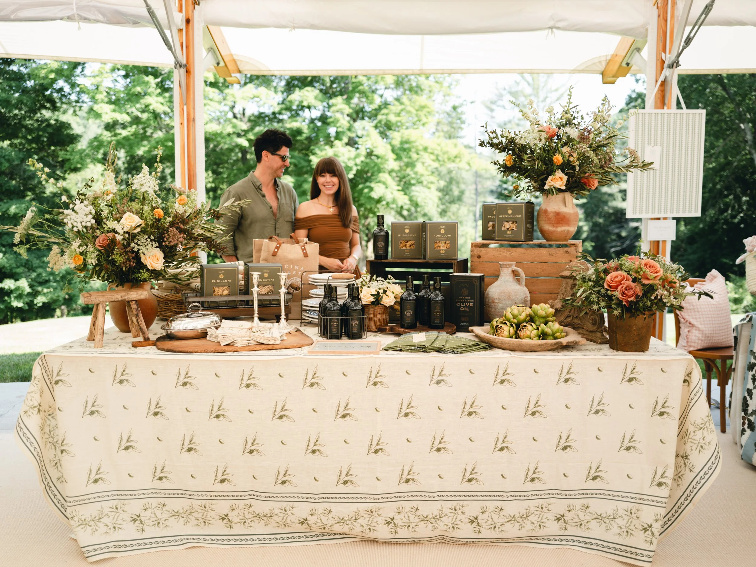 A man and a woman stand behind a table with floral arrangements and food products at an outdoor event under a white tent.