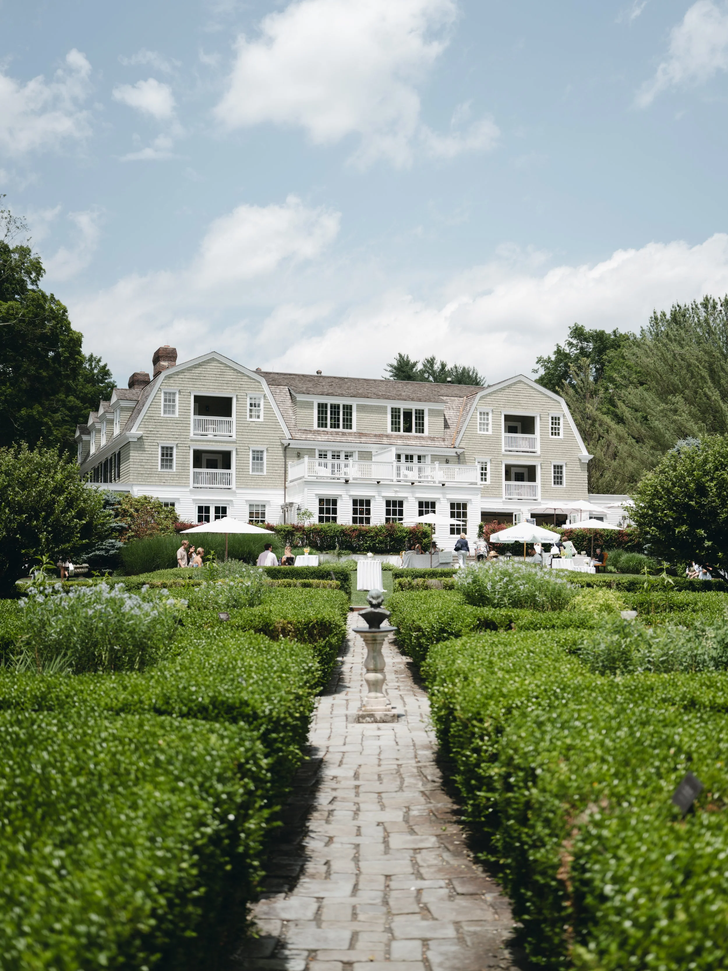 A large white house with multiple balconies, surrounded by a garden with hedges, trees, and a brick pathway leading to the house. There are people gathered outside under umbrellas.