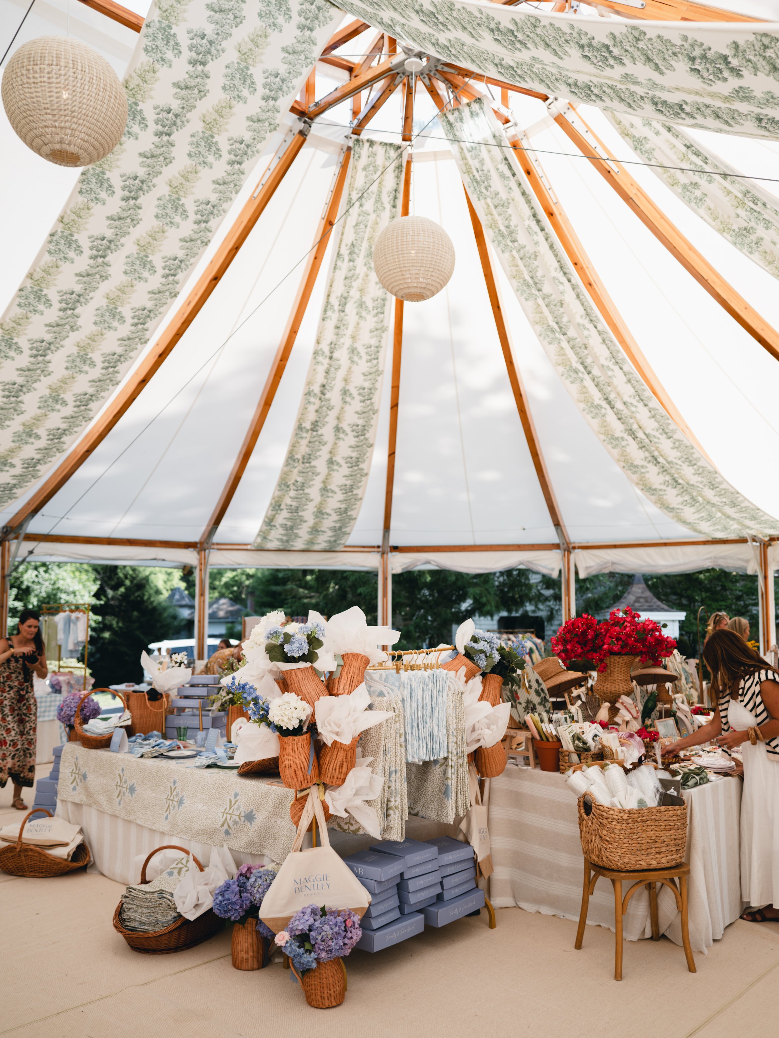 Outdoor market stall decorated with white and green fabric, displaying flowers, handbags, and various accessories, with people browsing.
