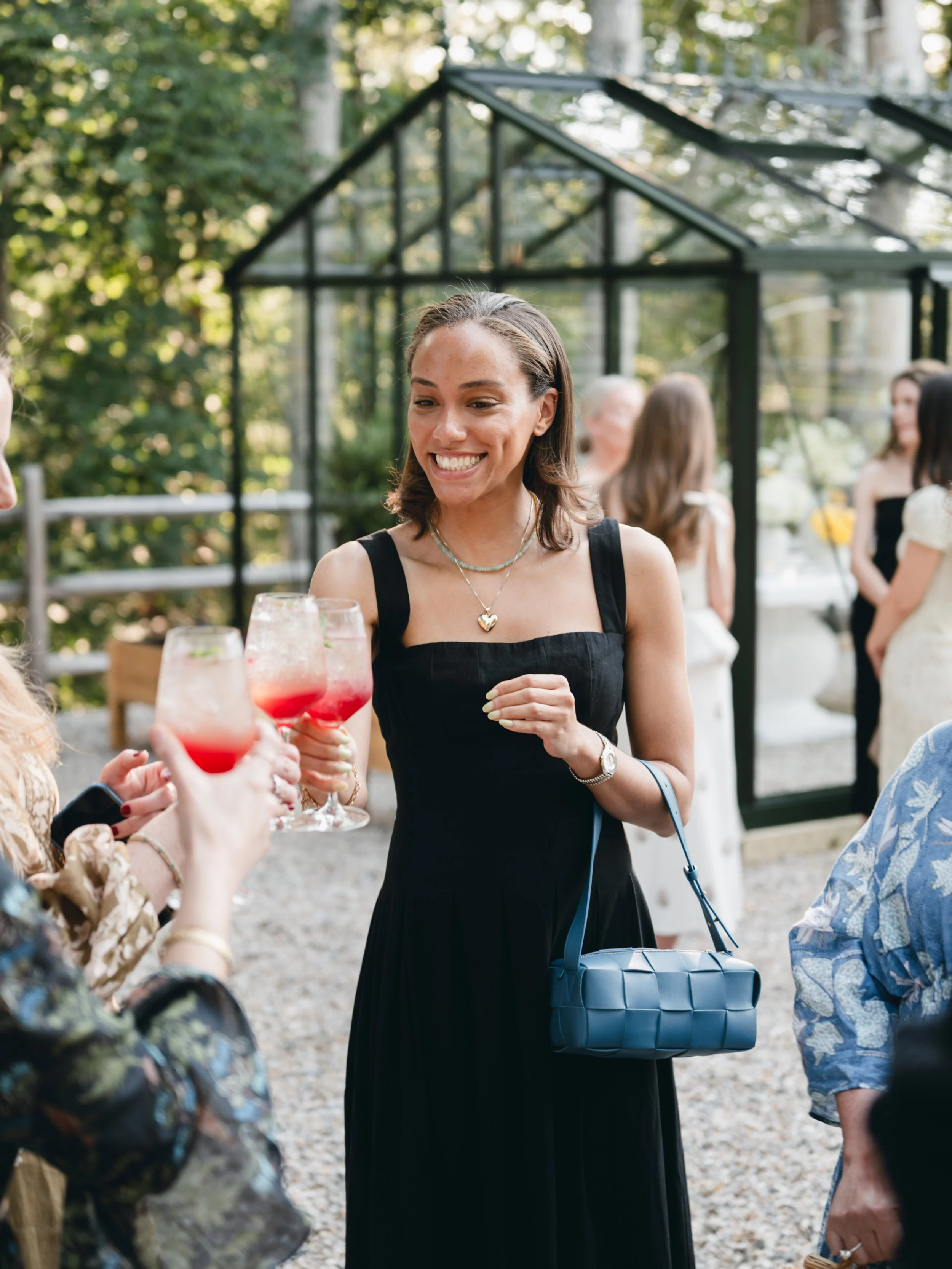 A woman wearing a black dress, jewelry, and carrying a blue purse, smiling as she holds a glass of pink drink during an outdoor gathering with other people in the background near a glass greenhouse surrounded by trees.