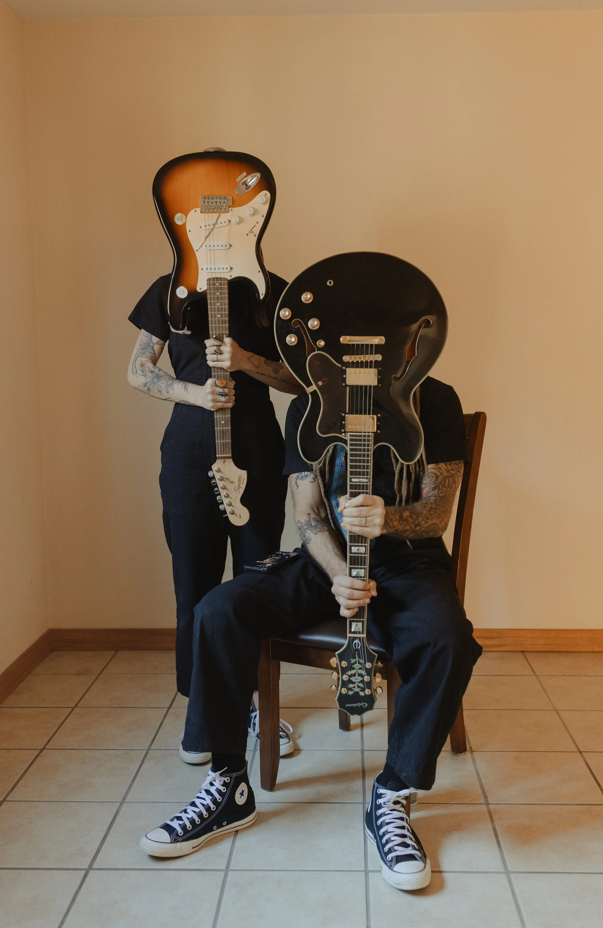 Two people with guitars for heads, one standing and one sitting, in a room with beige walls and tiled floor.
