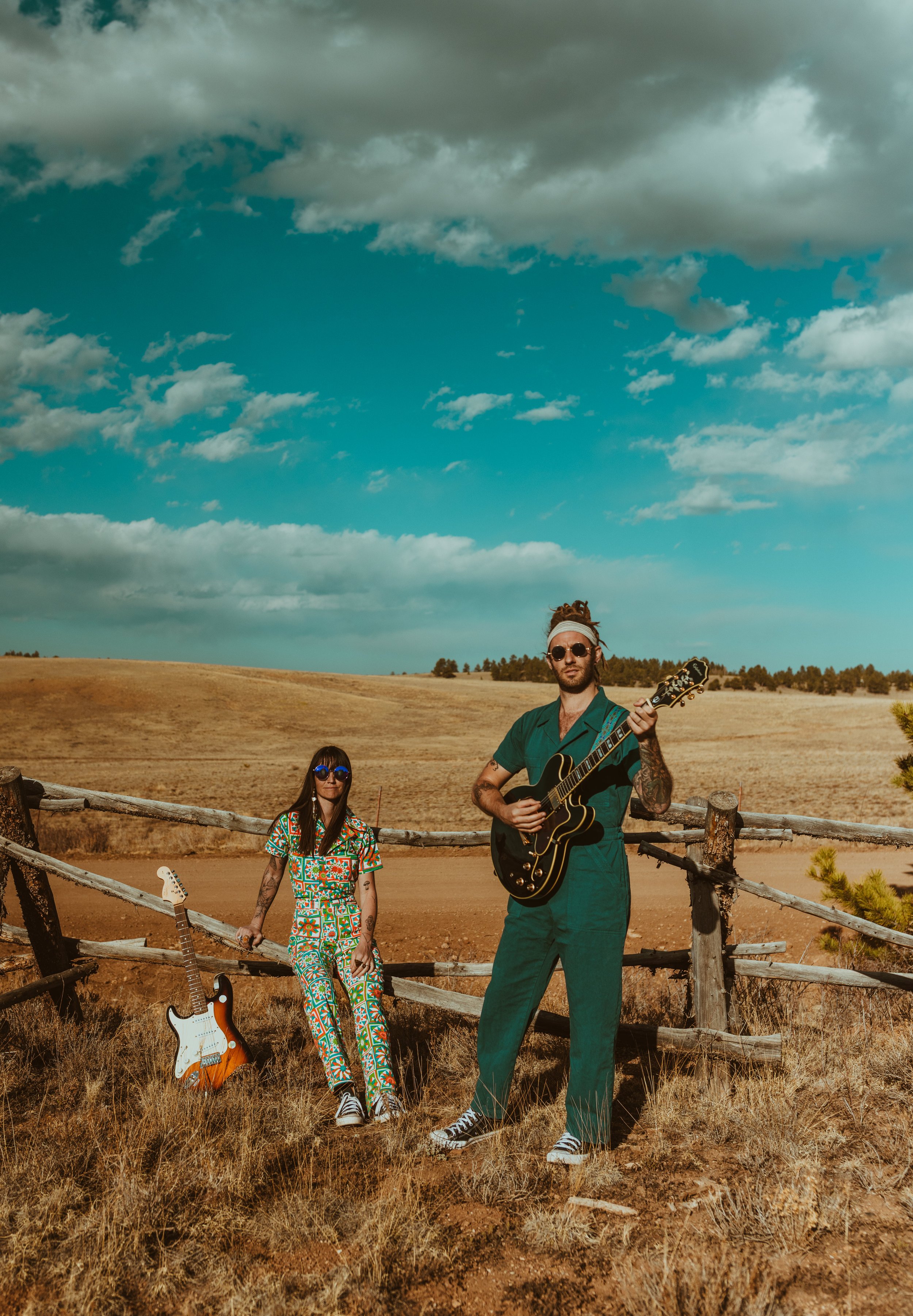 Two people standing outdoors in a rural landscape with a blue sky and clouds. One person is holding a guitar, and there is an electric guitar on the ground nearby.