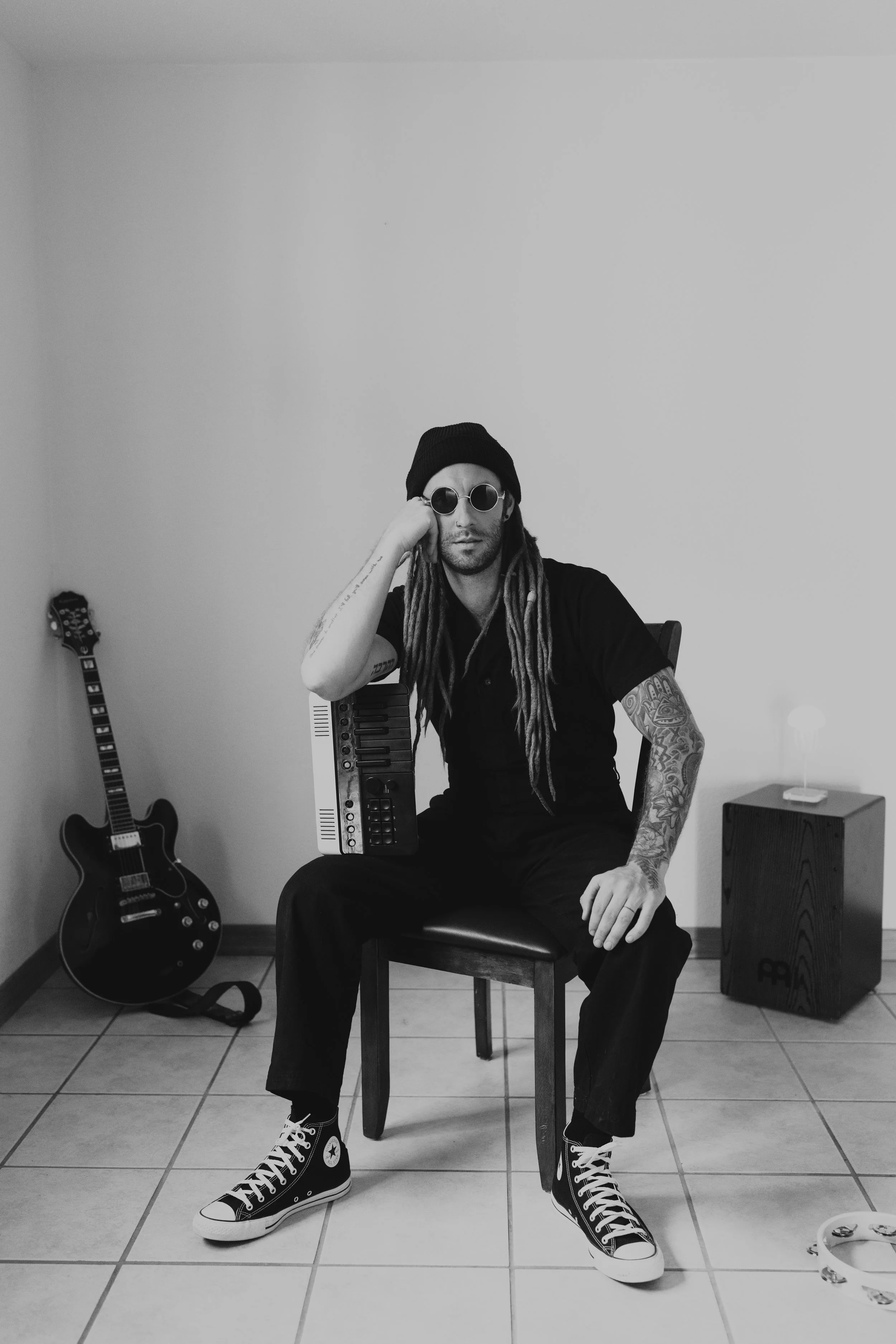 Man with long dreadlocks, sunglasses, beanie, and tattoos sitting on a chair in a music room with a guitar, keyboard, and cajón drum.