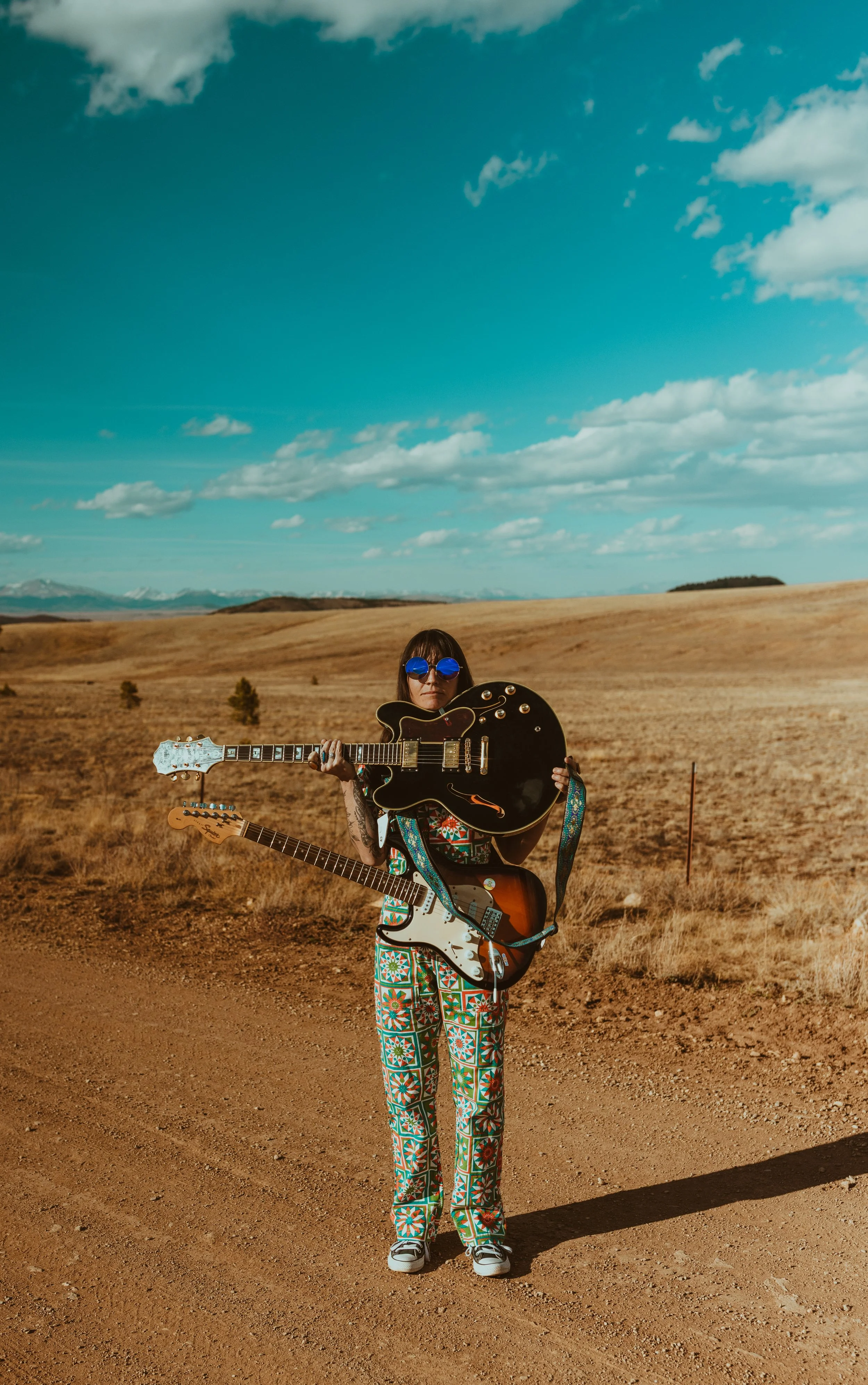 A woman standing on a dirt path in an open landscape holding two guitars, wearing colorful patterned pants, sunglasses, and a sleeveless top, under a partly cloudy sky.
