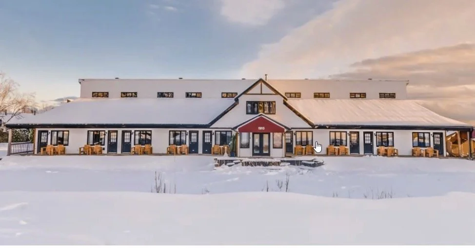 Un grand bâtiment en bois blanc avec un toit enneigé, des fenêtres sombres, et un porche à l'entrée, sous un ciel partiellement nuageux au coucher du soleil.