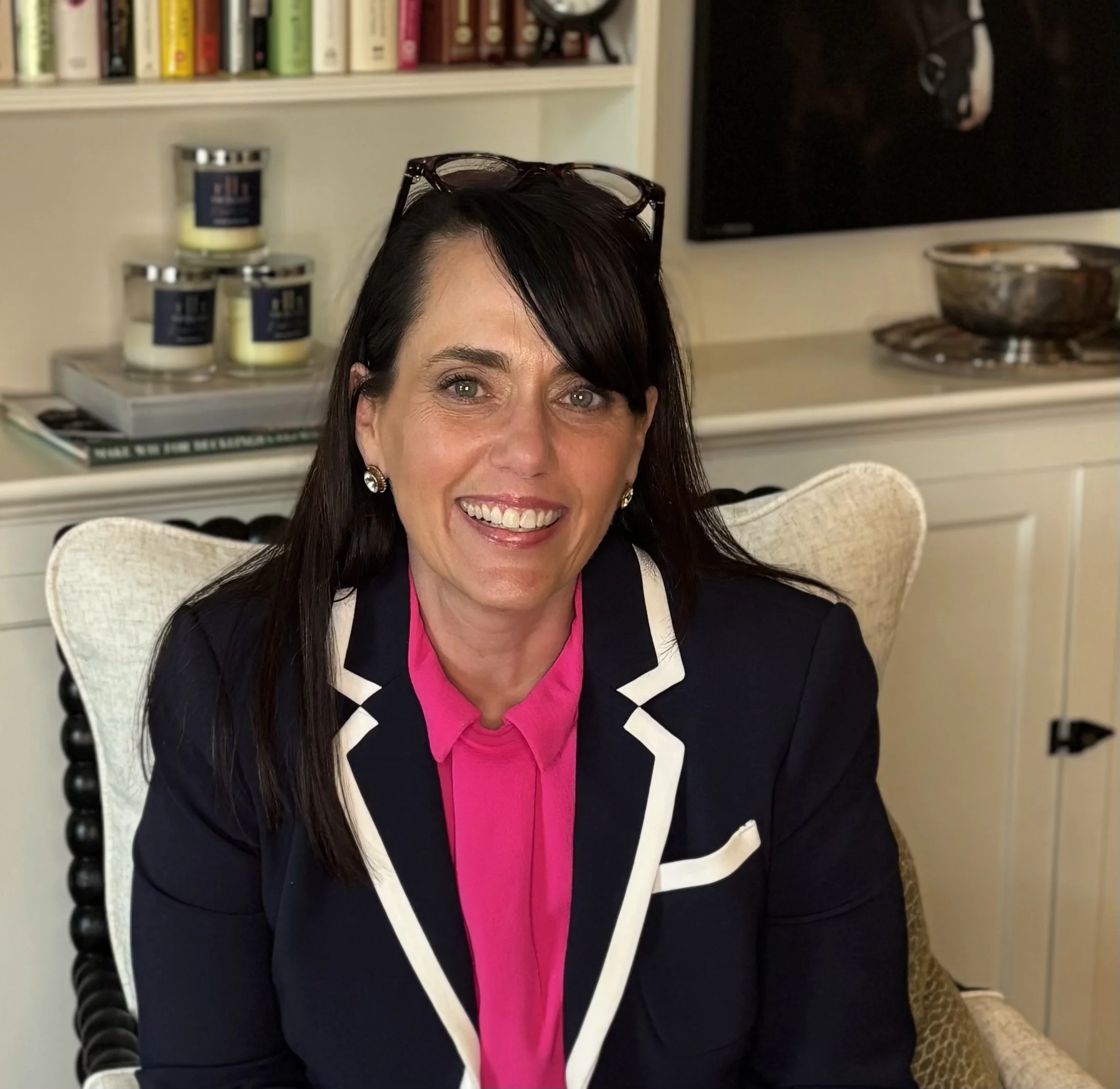 A smiling woman sitting in a chair at a desk, wearing a dark blazer with white trim and a bright pink blouse, with glasses resting on her head.