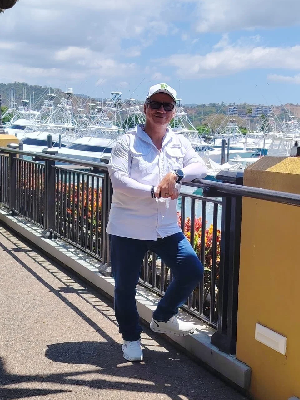 Un hombre con ropa blanca, gafas, gorra y sneakers blancos, posa frente a un muelle con varios barcos en el mar, con cielo parcialmente nublado.