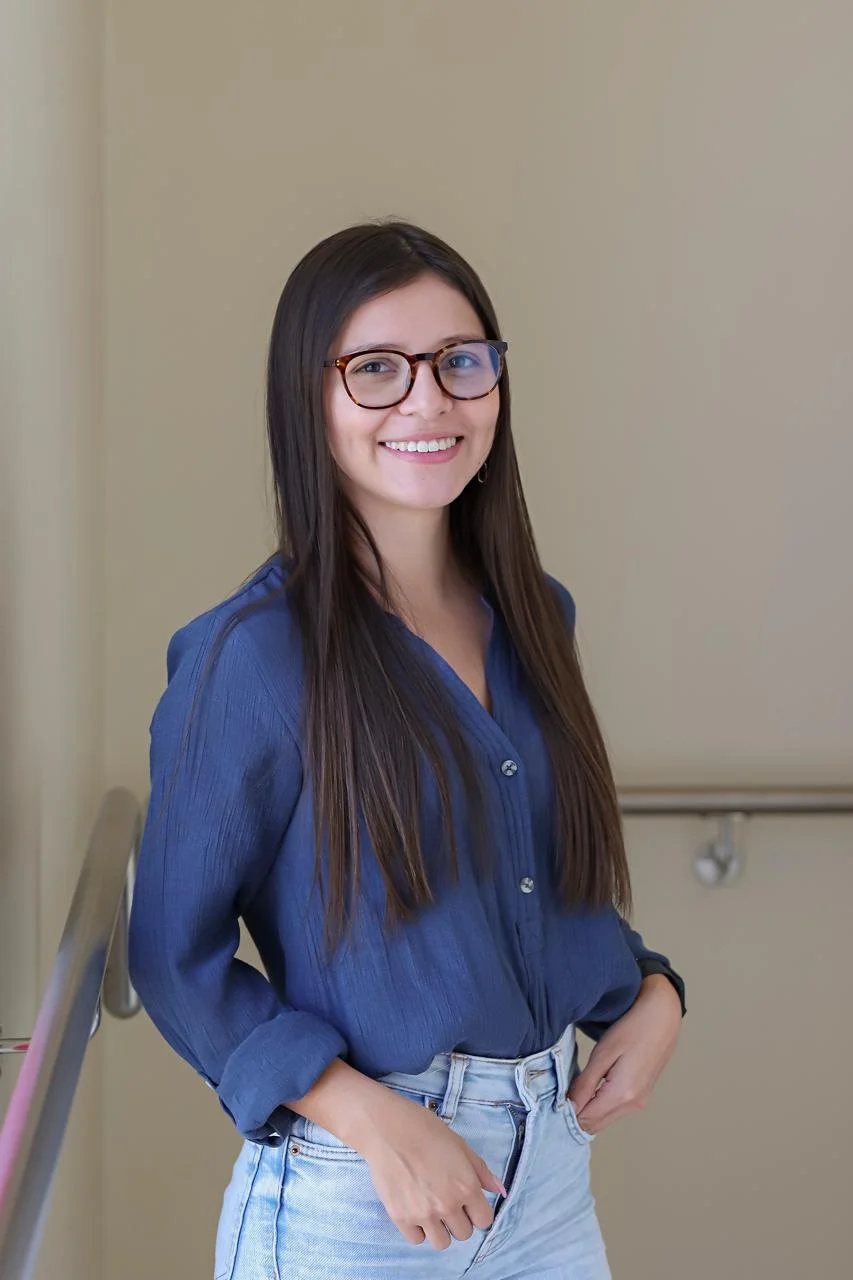 Joven mujer con gafas, cabello largo y oscuro, vestida con blusa azul y jeans, sonriendo.