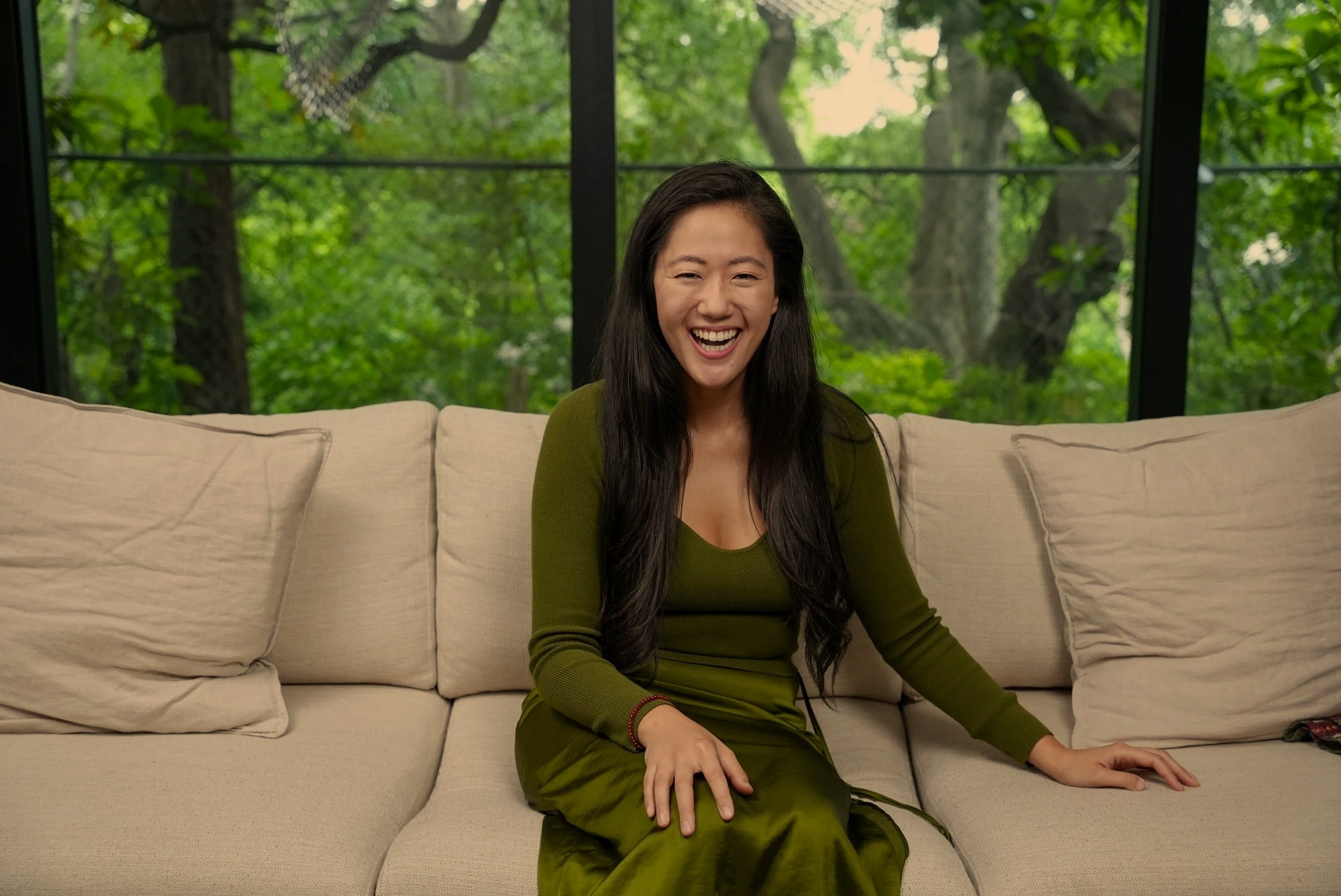 A woman with long black hair, wearing a green long-sleeve dress, sitting on a beige couch, smiling and laughing, in front of a window with green trees outside.