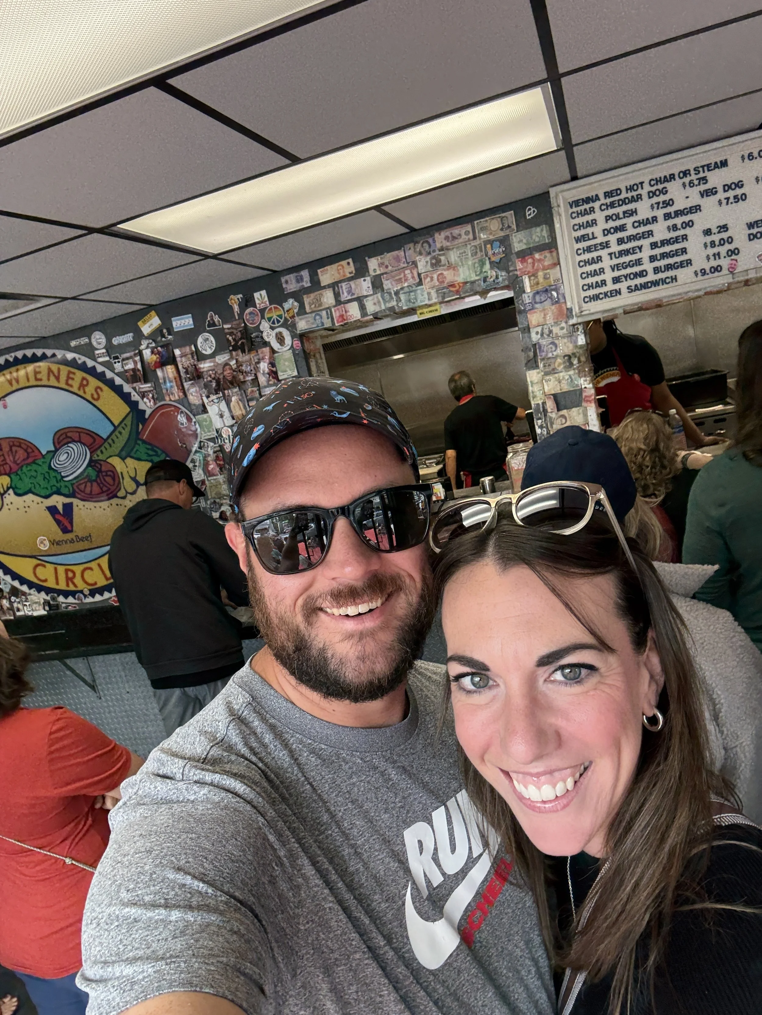 The owner and his wife smiling at a famous Chicago hot dog restaurant, with a busy kitchen in the background and a menu above them listing various hot dogs and burgers.