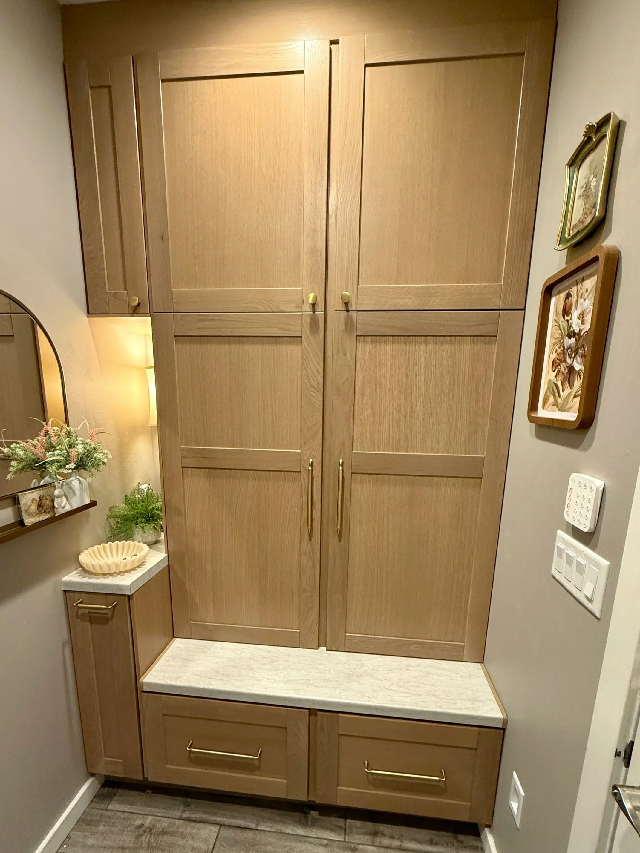 Wooden cabinet with two doors and gold handles, a small shelf with a flower arrangement on a ledge, and colorful wall art depicting flowers on the right wall. Light switch and electrical outlets are visible on the gray wall.
