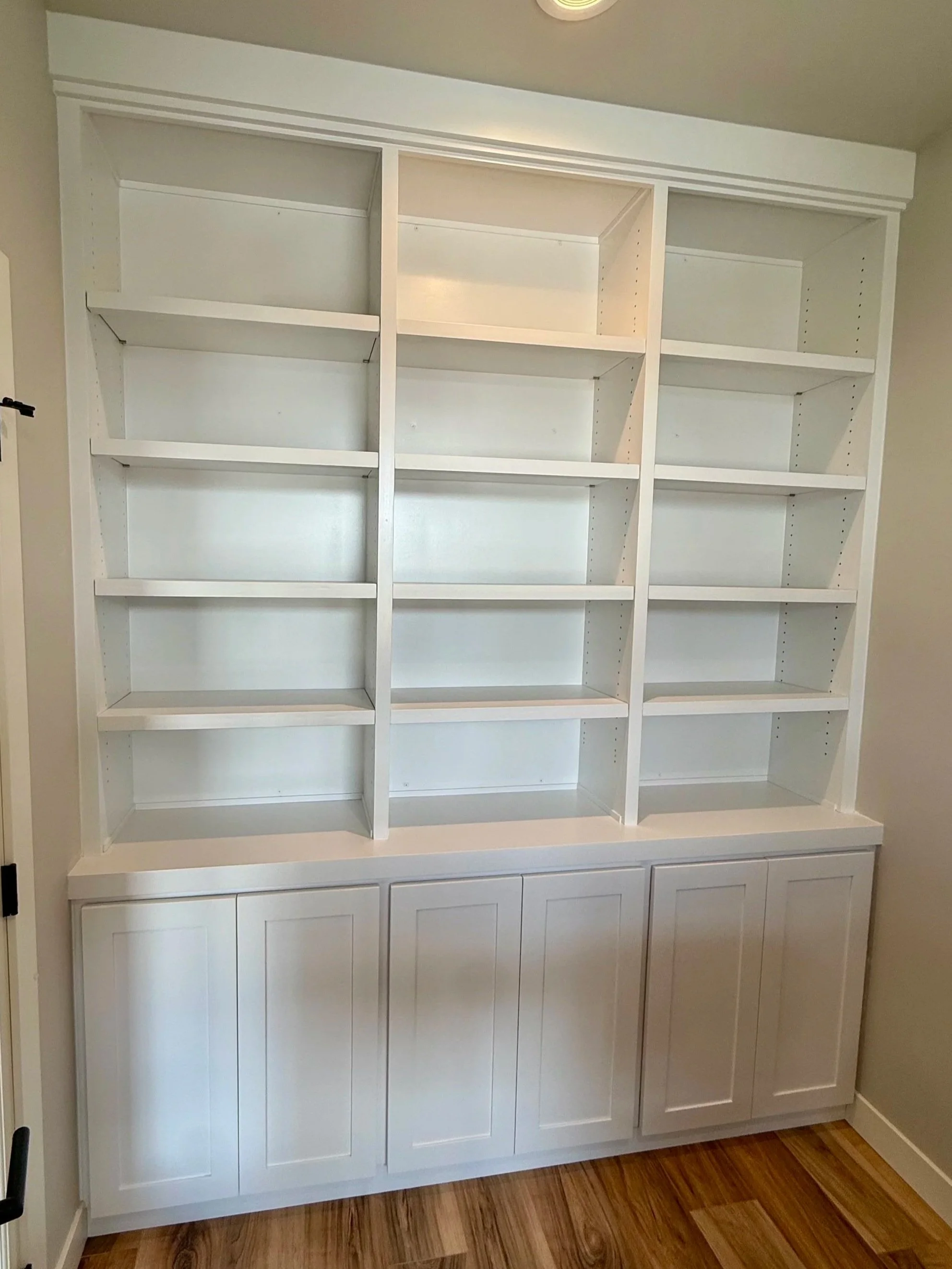 A large white built-in bookshelf with multiple open shelves and cabinet doors at the bottom, located against a beige wall on a hardwood floor.