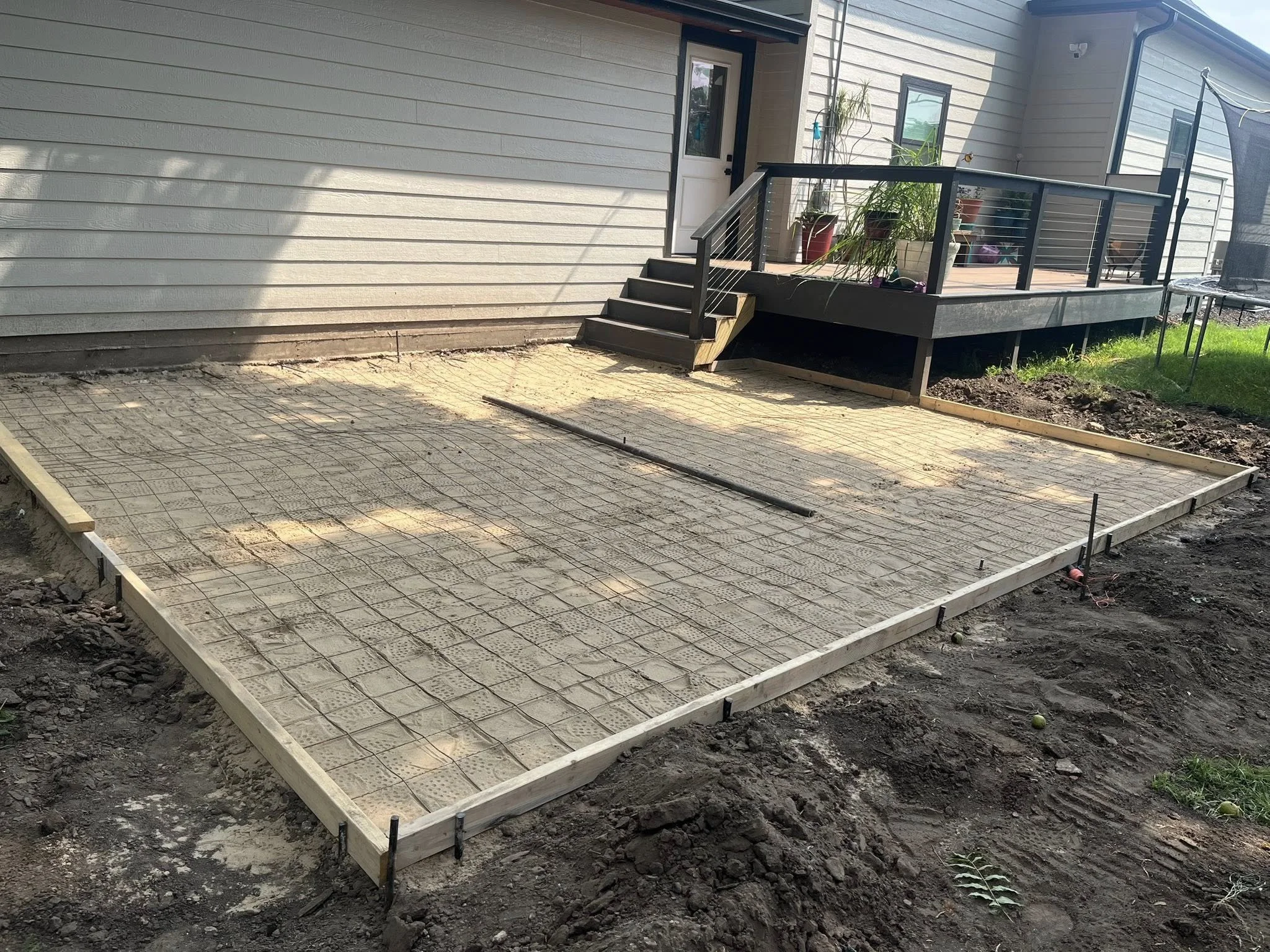 A construction site in a backyard with a wooden frame and rebar grid, preparing for concrete pouring, next to a house with a deck and potted plants.